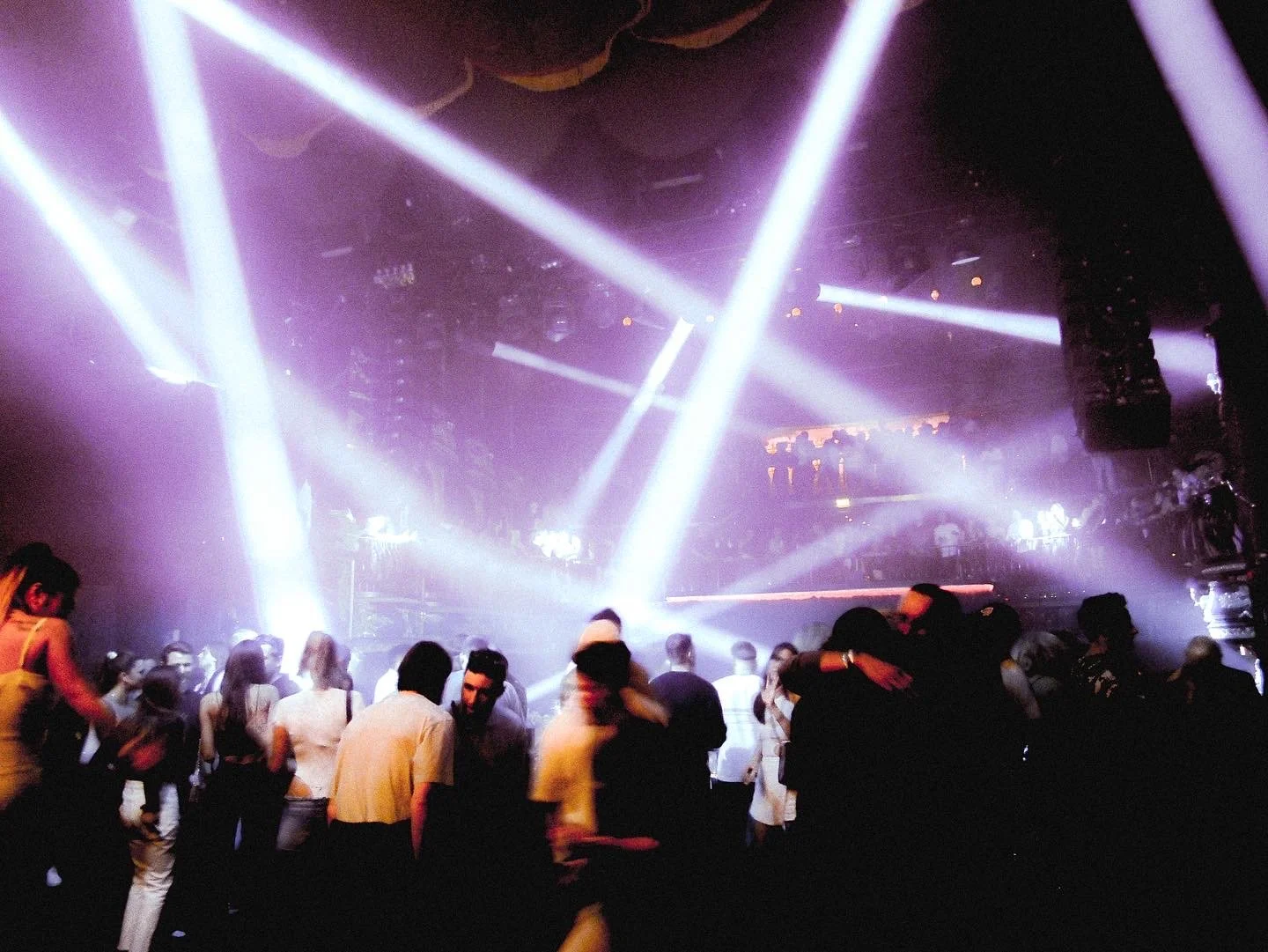 People dancing in a nightclub with colorful lights and fog, illuminated by bright stage lighting.