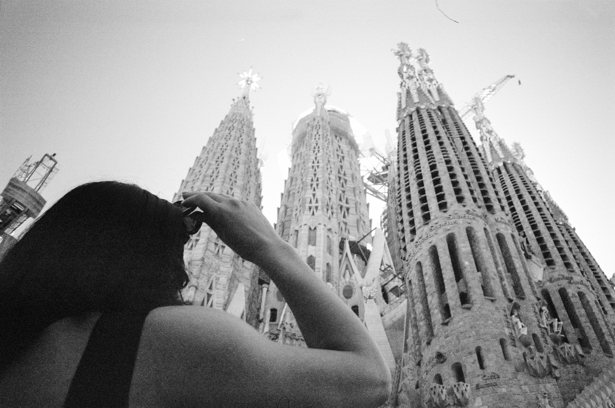 Black and white photo taken from below of Person holding hands with another person, with Sagrada Familia church under construction in the background in Barcelona, Spain.