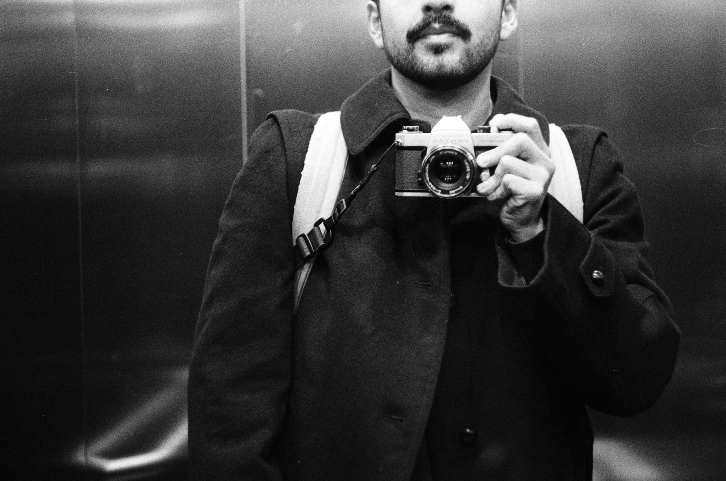 A man taking a selfie in an elevator, holding a vintage camera up to his face. He is wearing a dark jacket and has a backpack over his shoulder.
