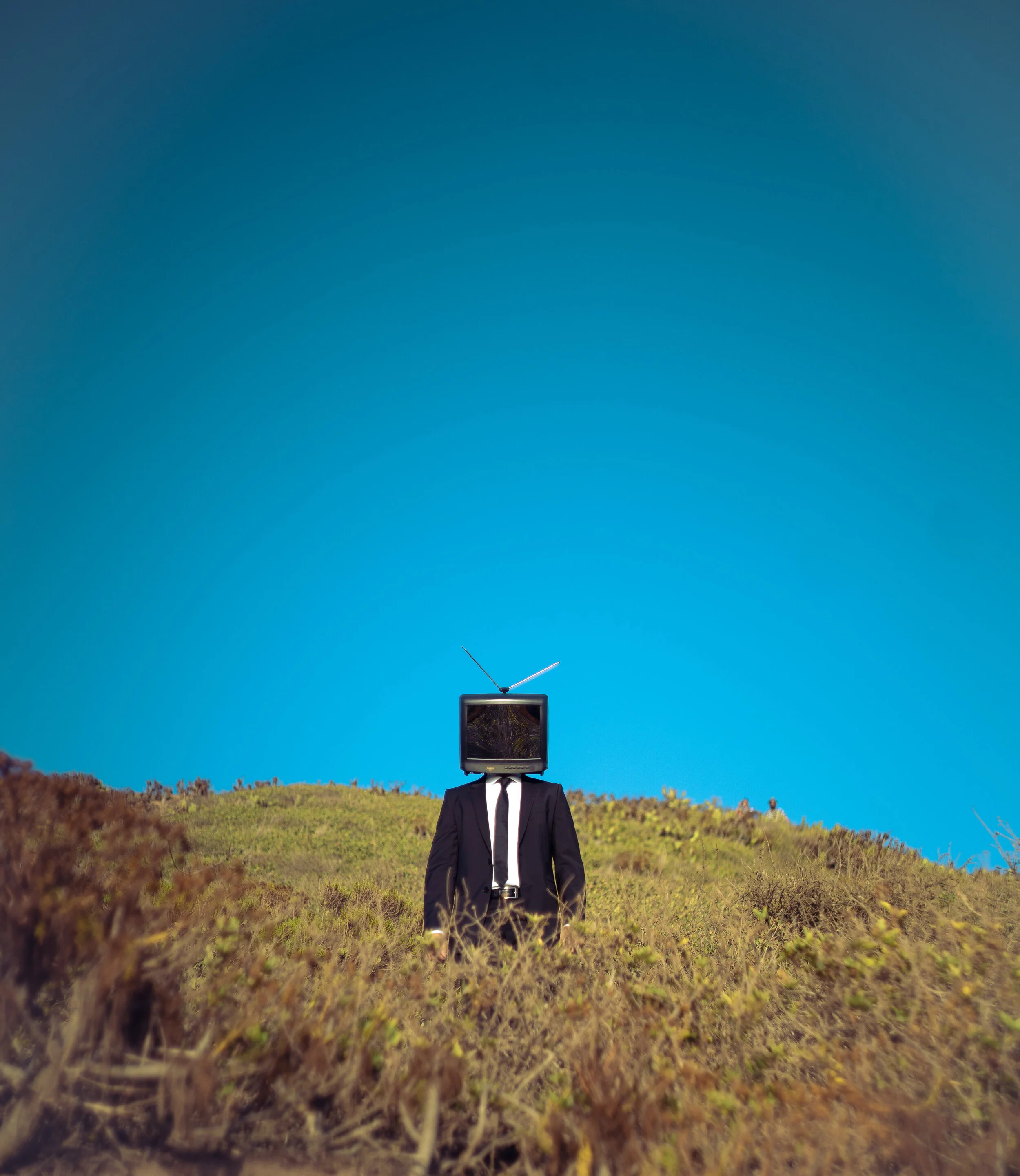 A person in a suit with a television for a head, standing on a grassy hill under a clear blue sky, with an antenna on top of the television.
