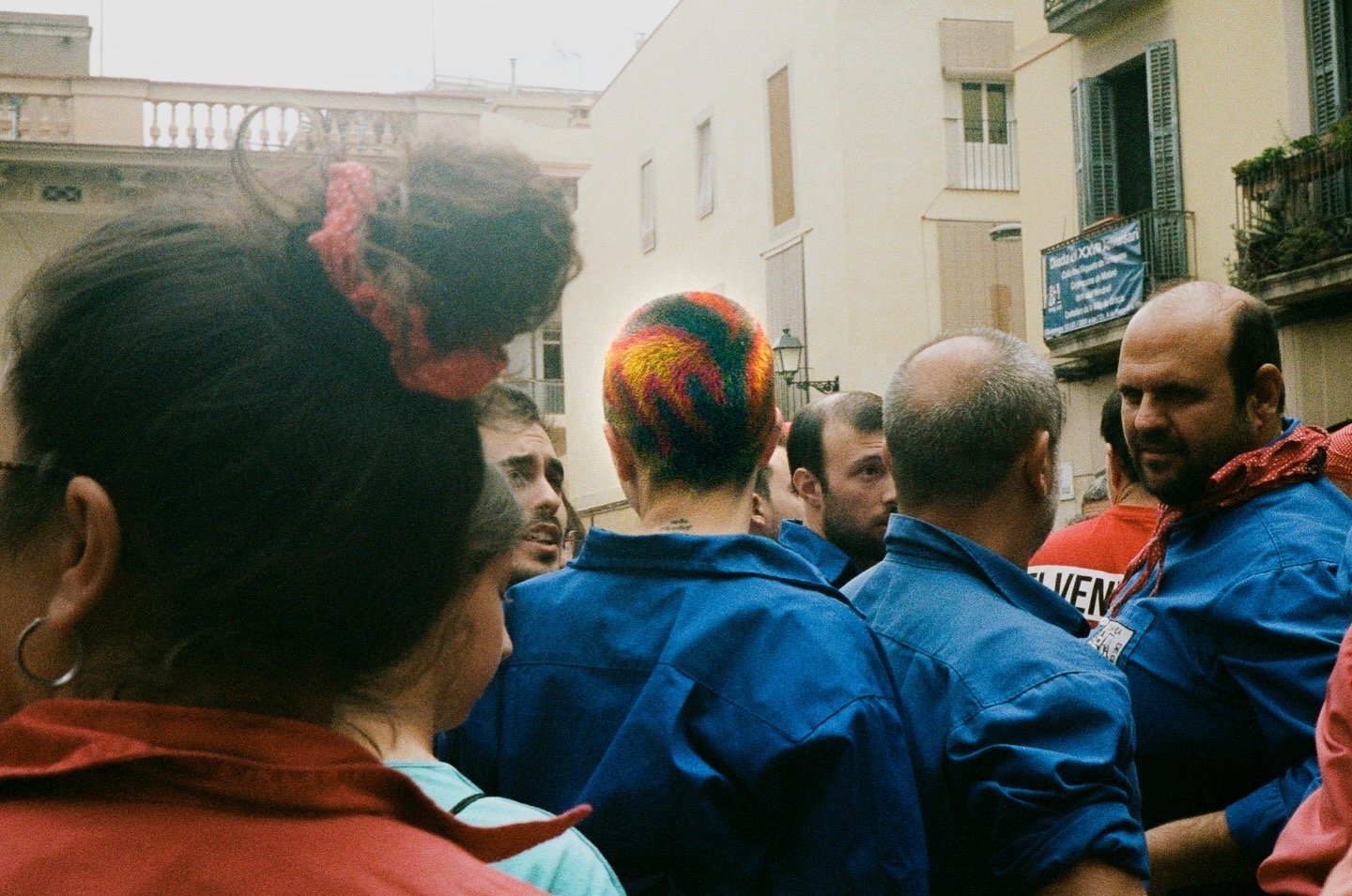 A group of people, some with colorful hair, gathered outdoors in an urban area with buildings and balconies, engaging in conversation.