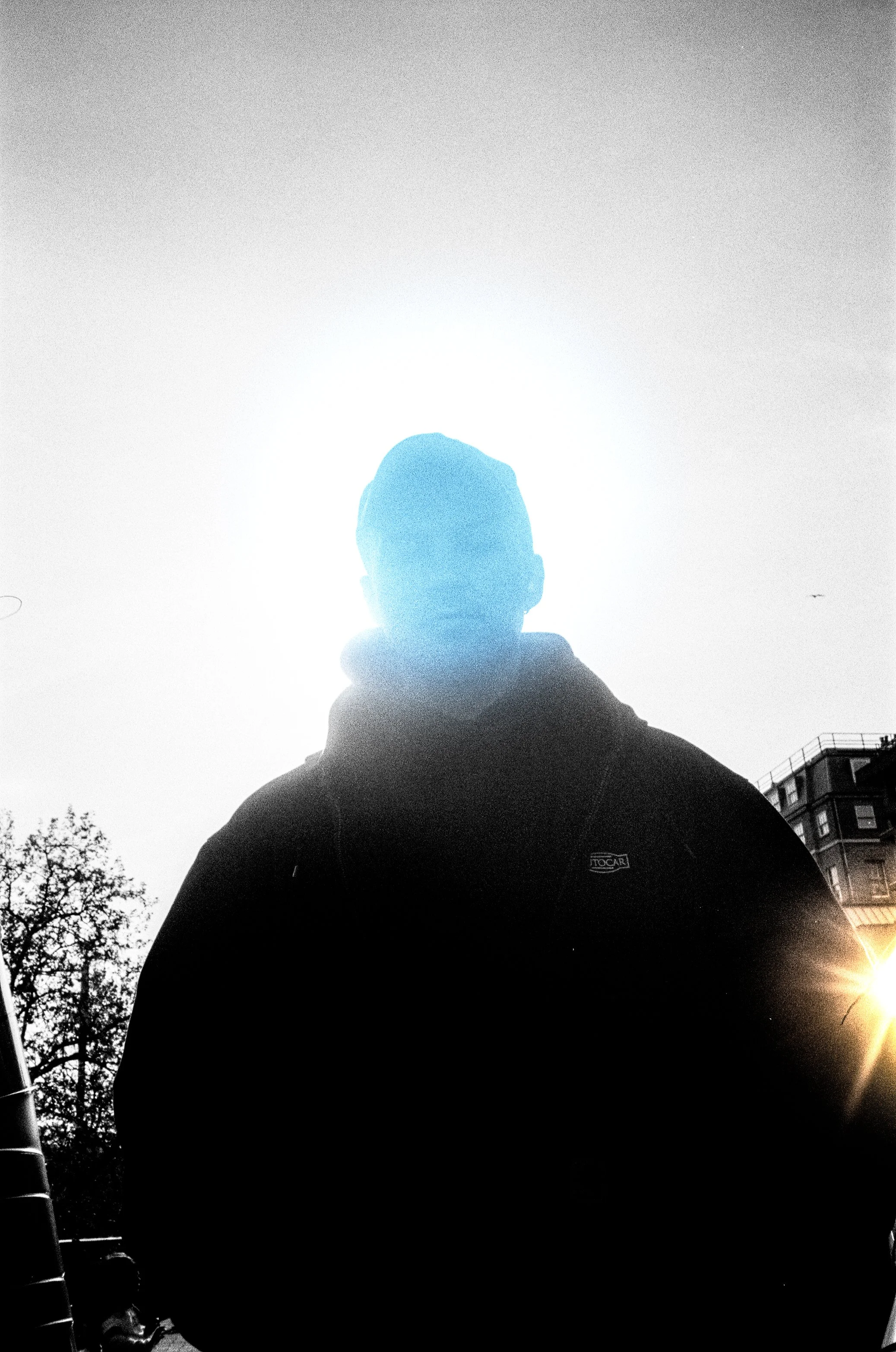 A silhouette of a person seen from behind, wearing a hoodie, standing outdoors in front of a bright sky with the sun shining near the horizon, with trees and a building in the background.