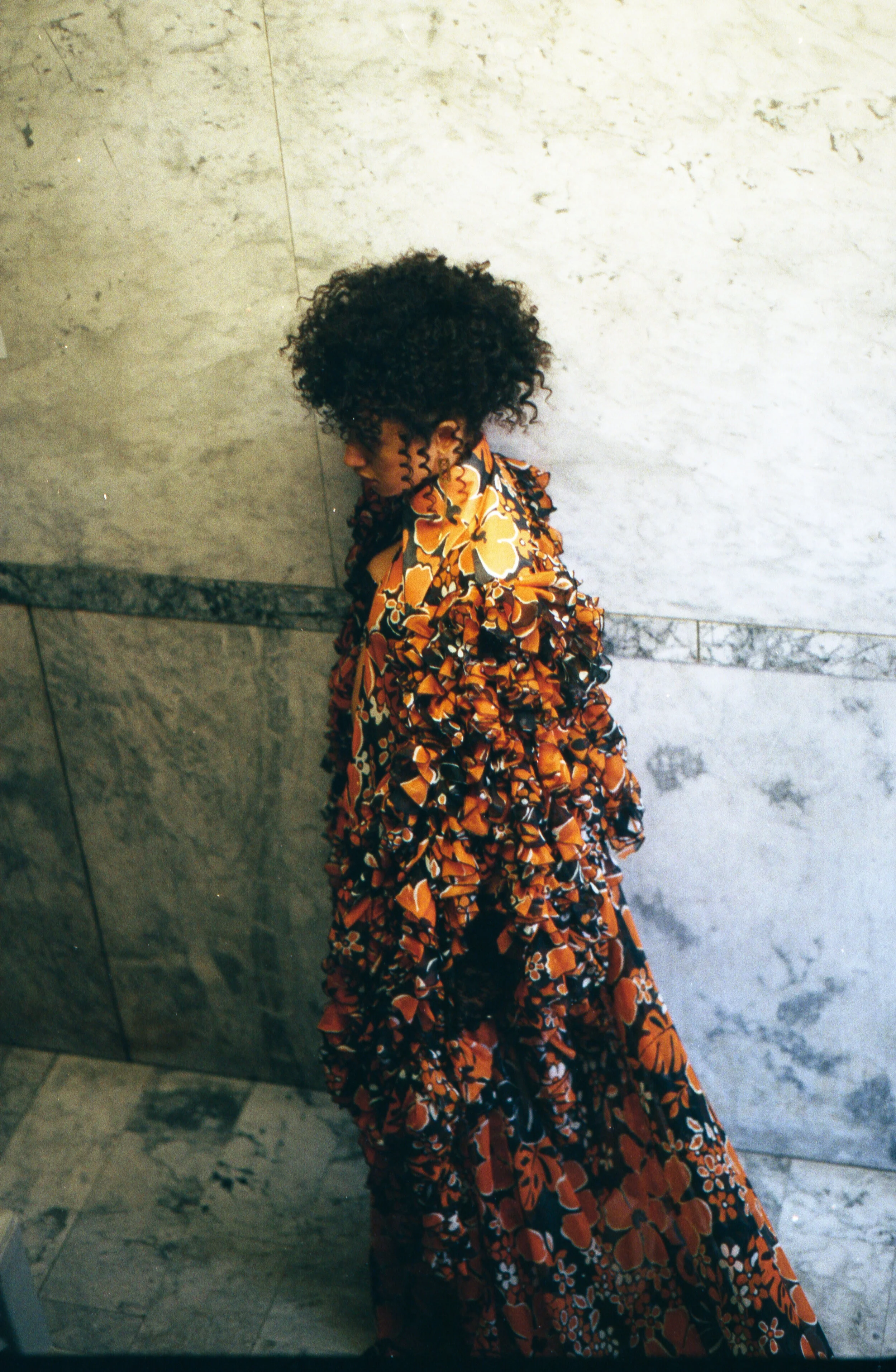A woman with curly hair wearing a long, orange floral dress stands against a gray marble wall. She looks down with a serious expression.