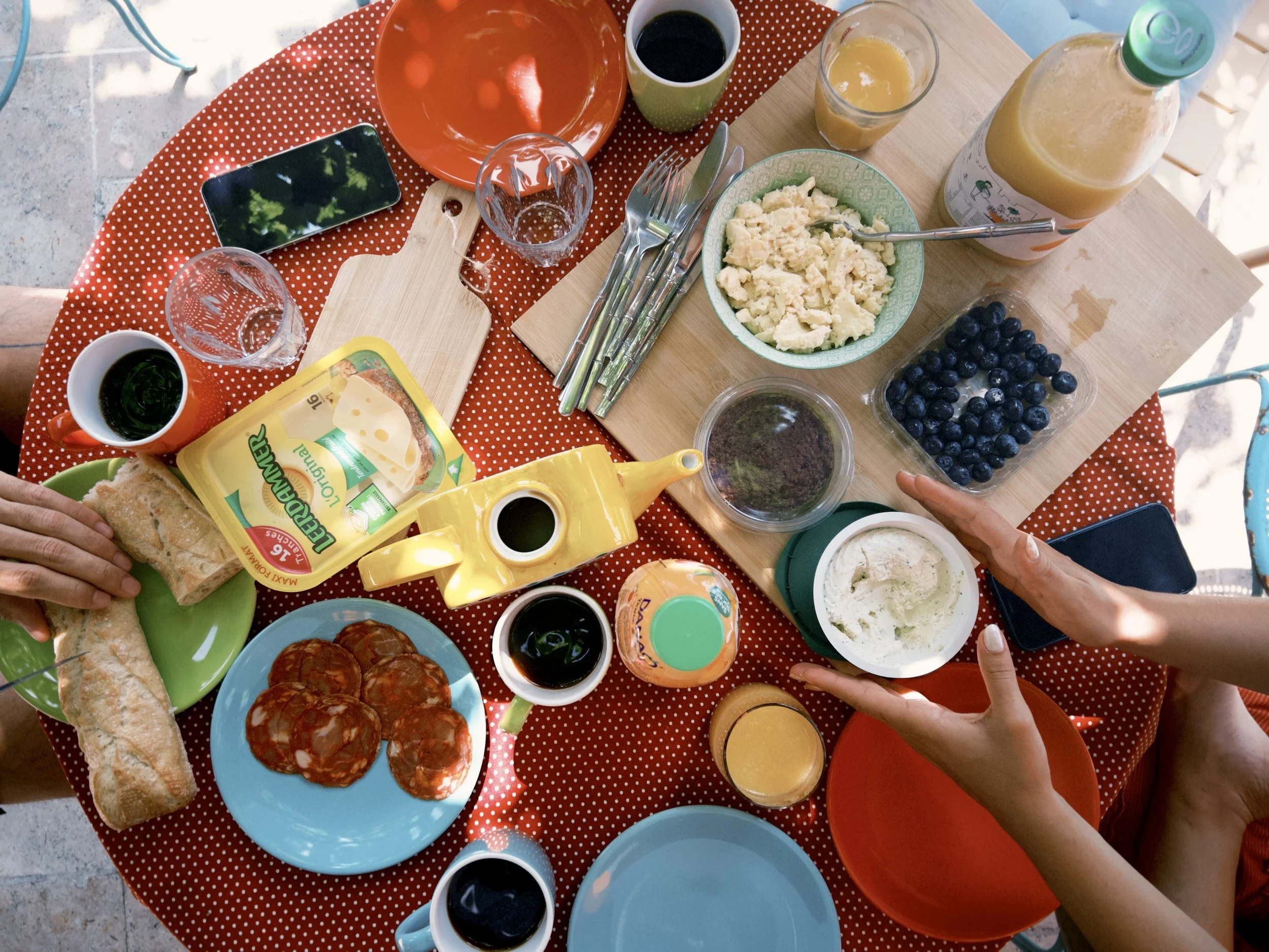 A table set for breakfast with various foods including blueberries, scrambled eggs, slices of bread, slices of pepperoni, and condiments. There are glasses of orange juice, coffee, and a bottle of orange drink. A person's hands are visible, one holdi