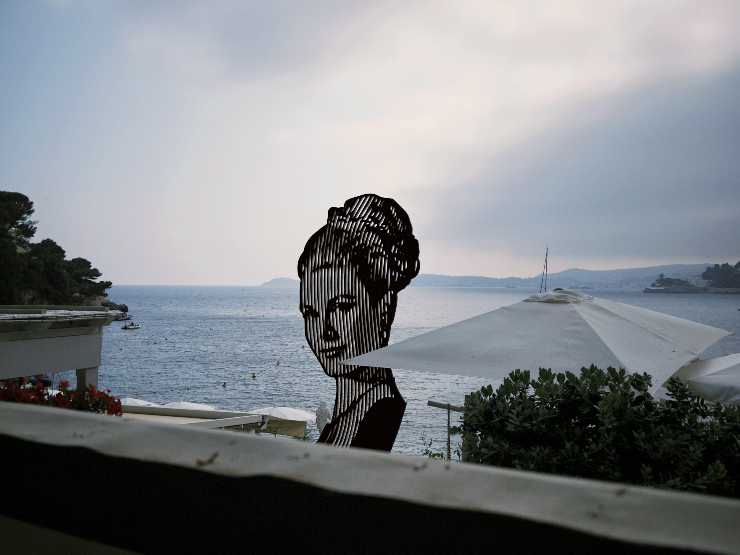 Silhouette of a woman’s profile made of metal against a waterfront view with trees, water, and a sailboat in the distance, under a partly cloudy sky.