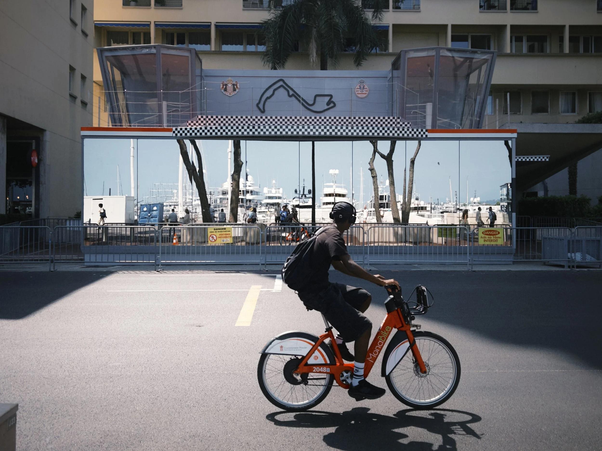 A person riding an orange and white bicycle labeled 'Mobike' on a city street with yachts docked at a marina in the background. The cyclist is wearing a black t-shirt, dark shorts, and a backpack.