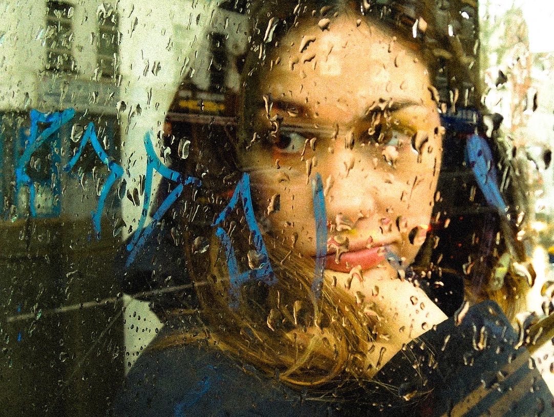 A woman looking through a rain-covered window with raindrops on the glass and blue writing on the window.