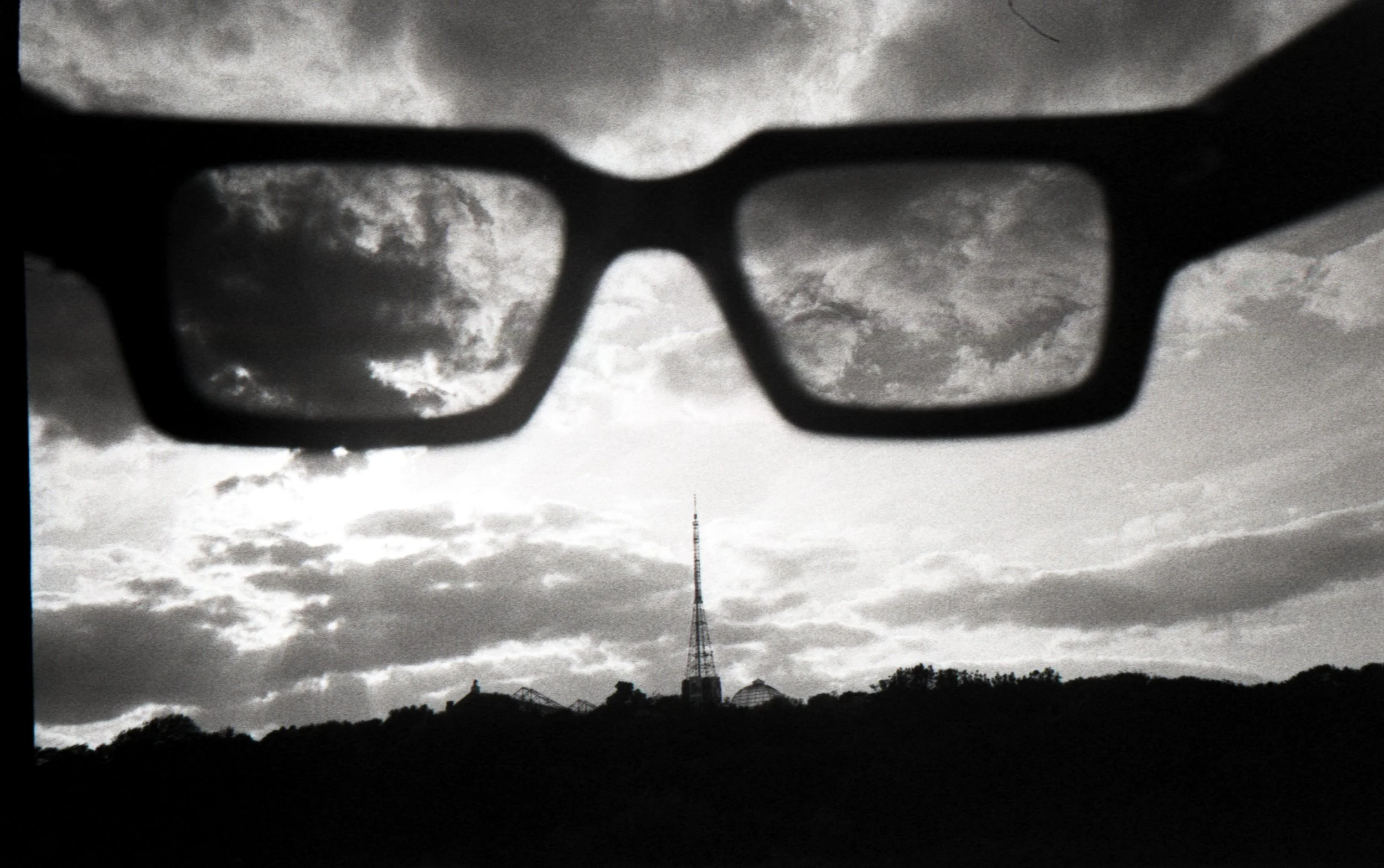 Black and white photo of sunglasses held up against the sky with clouds.
