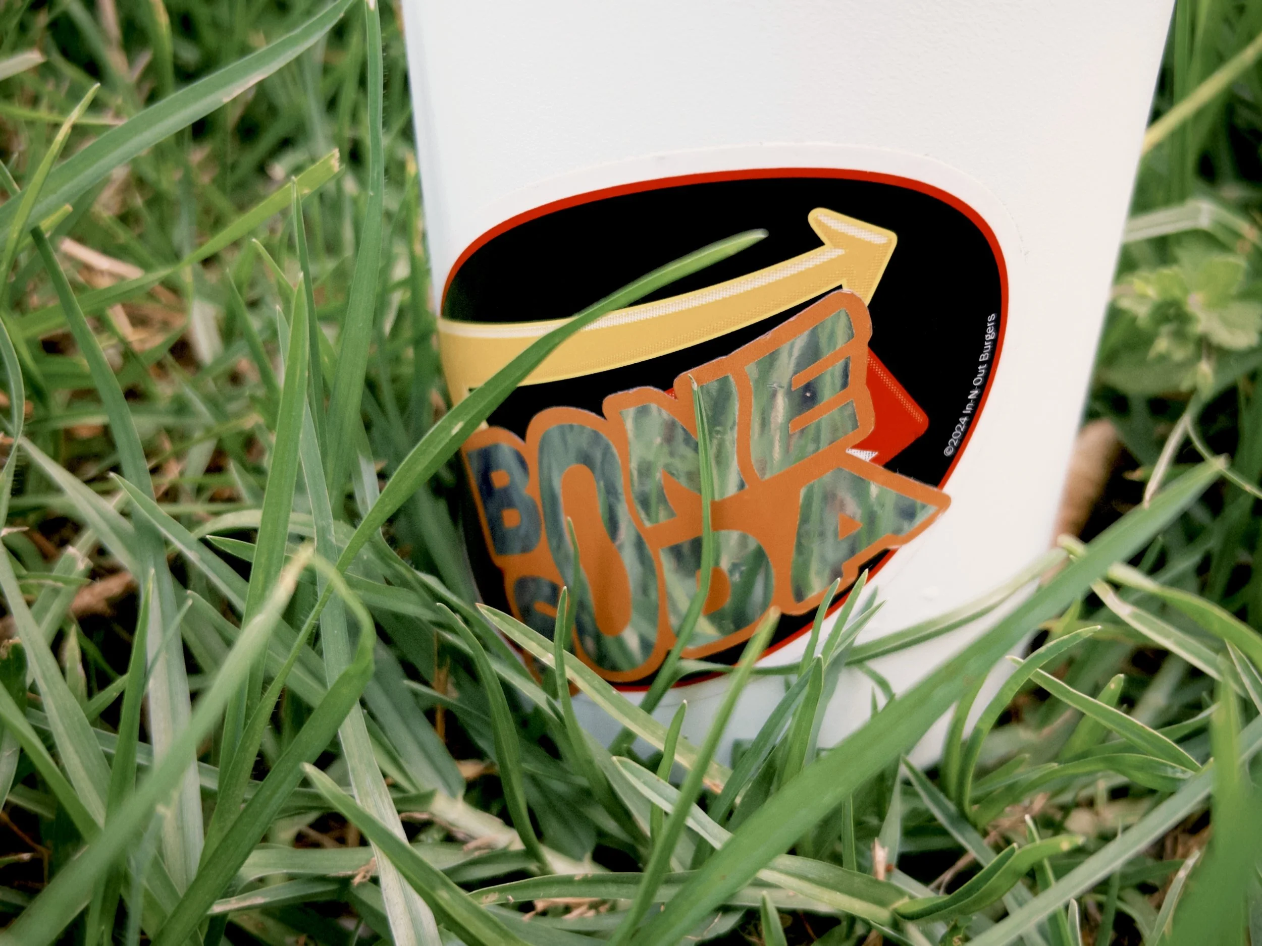 A close-up of a white cup with a partially visible logo, placed on grass blades. The logo features a stylized burger with an upward arrow.