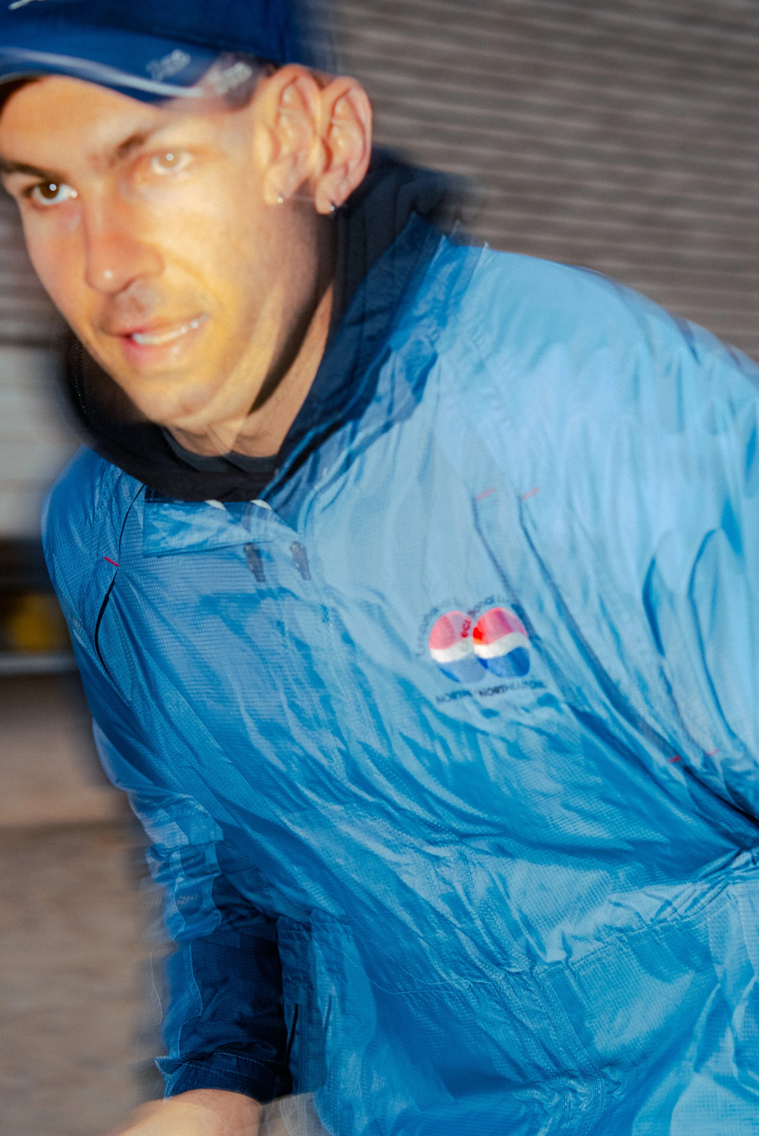 A man wearing a blue jacket with a Pepsi logo, a black hood, and a black cap, in motion, with a blurred background and a motion effect on the image.