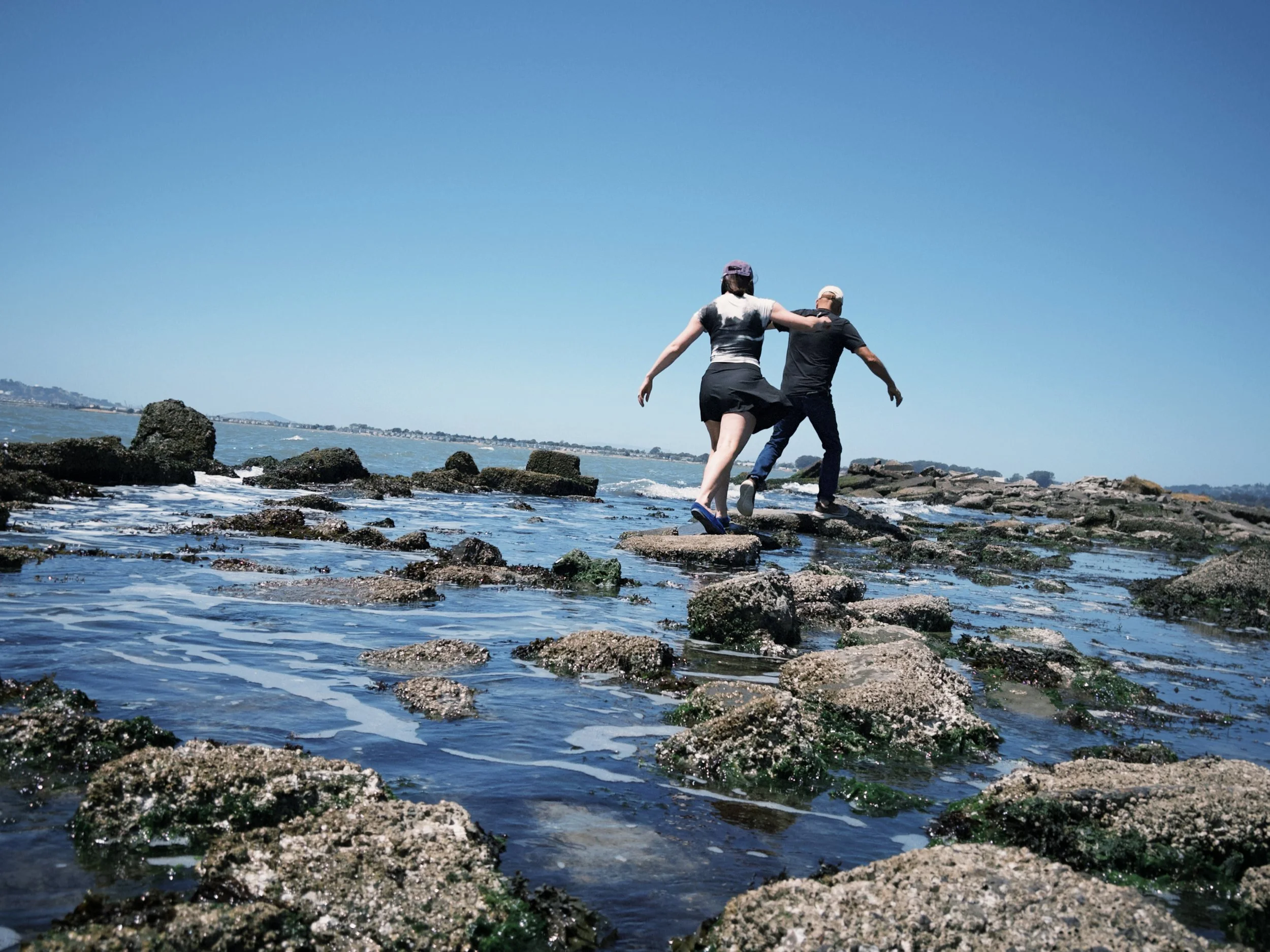 Two people walking on rocks by the shoreline with water and a distant landscape under a clear blue sky.