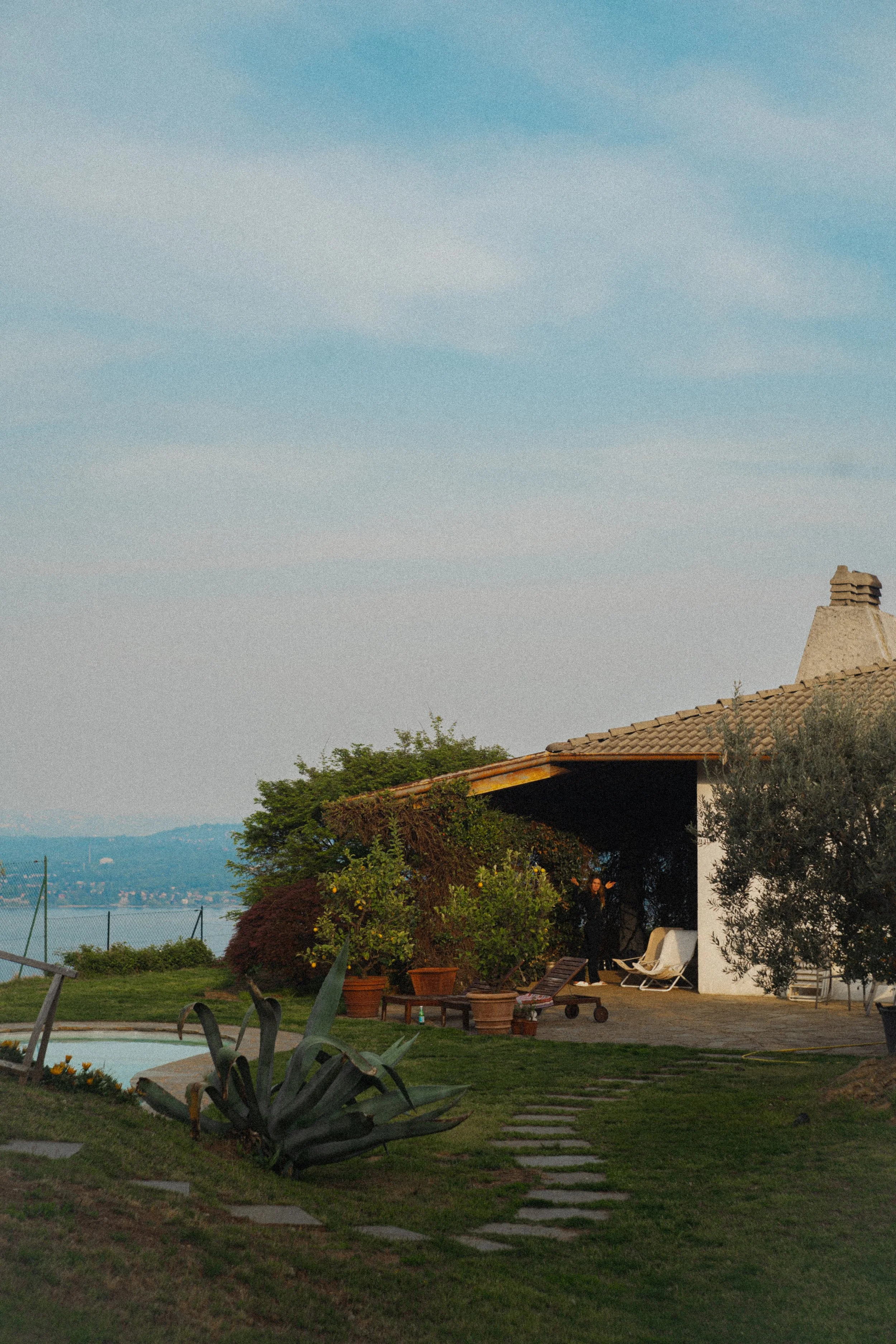 A backyard scene with a house that has a steep roof and chimney, surrounded by green bushes and trees, with a swimming pool and patio chairs in the yard, and a view of water and hills in the distance under a partly cloudy sky.