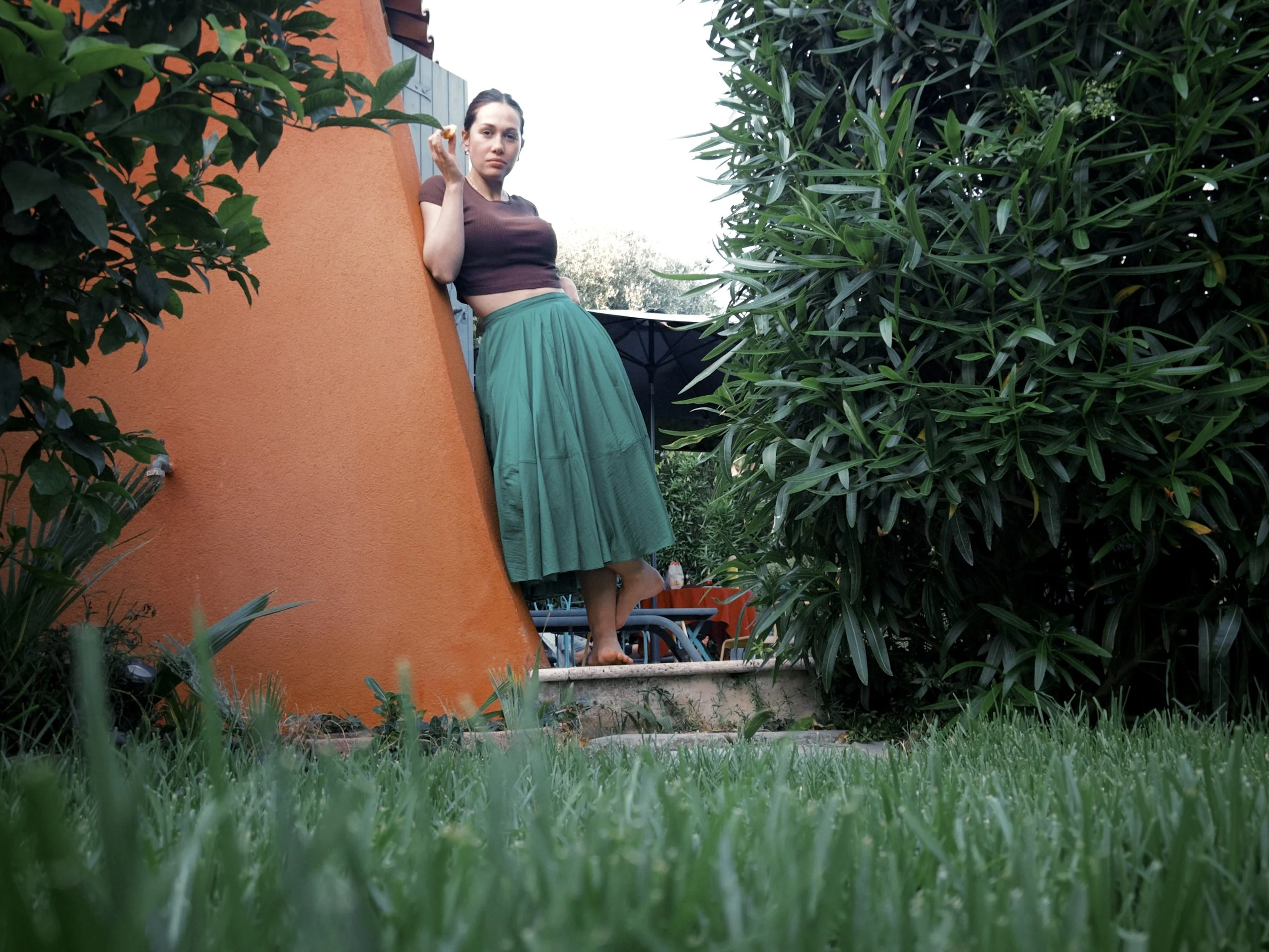 A woman in a brown crop top and green flowy skirt stands outside next to an orange wall, surrounded by lush green plants, holding a snack and looking at the camera.