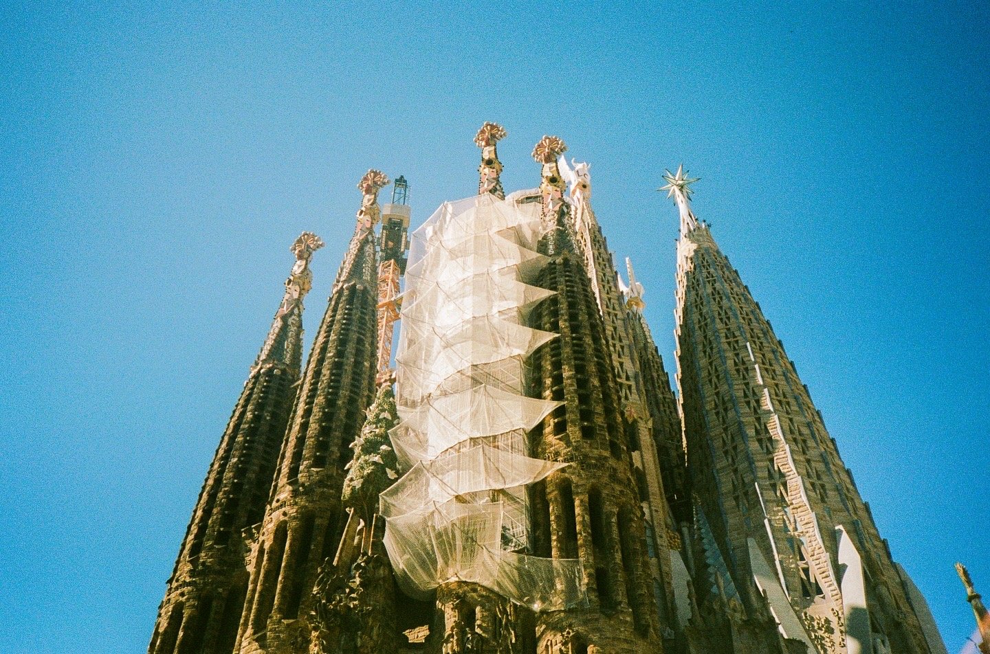 The Sagrada Família with a clear blue sky in the background, showing its intricate Gothic and Art Nouveau architecture, with spires and a construction scaffold.