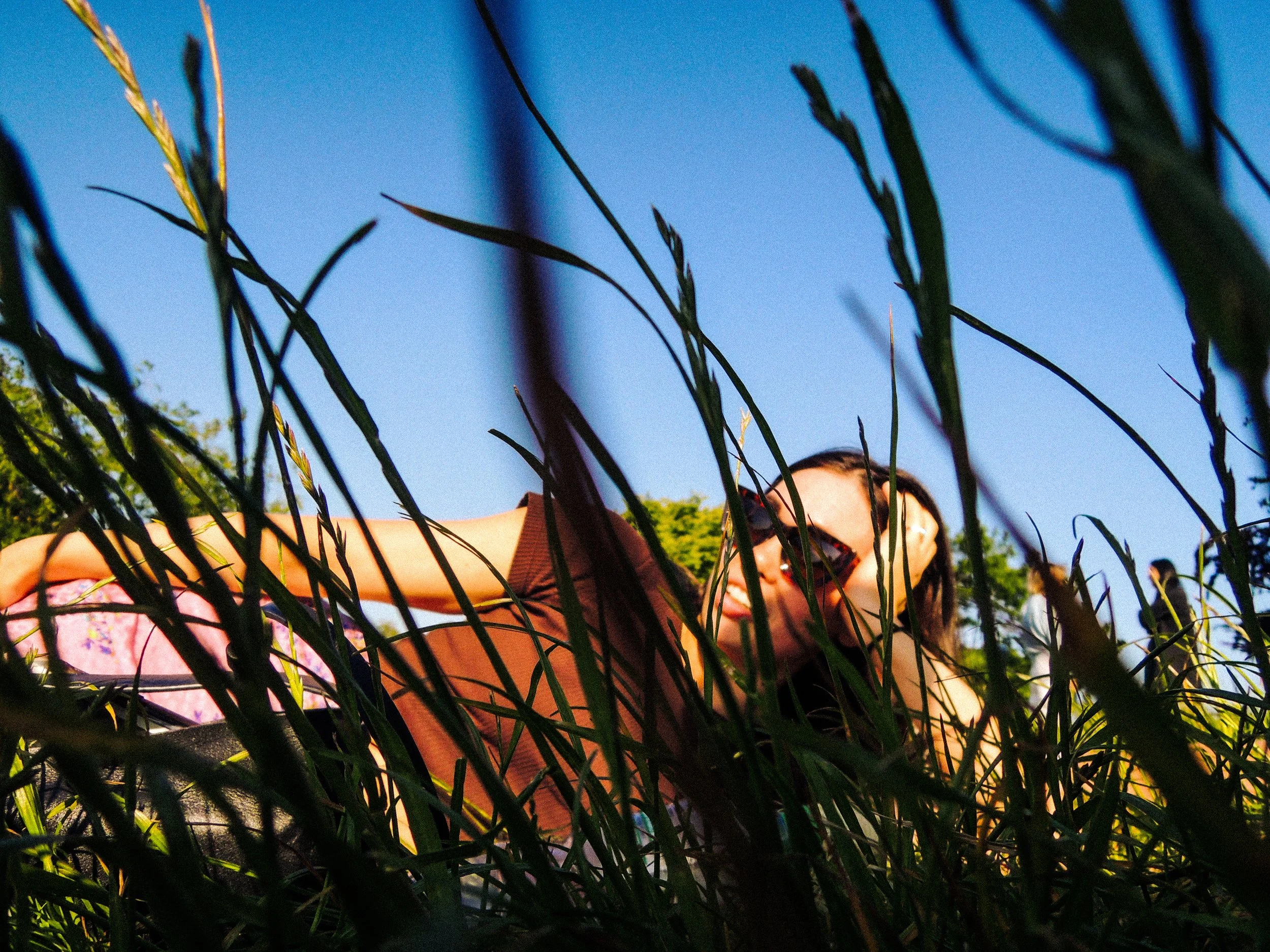A person with sunglasses and earrings lying on the grass among tall green grass with a clear blue sky in the background.