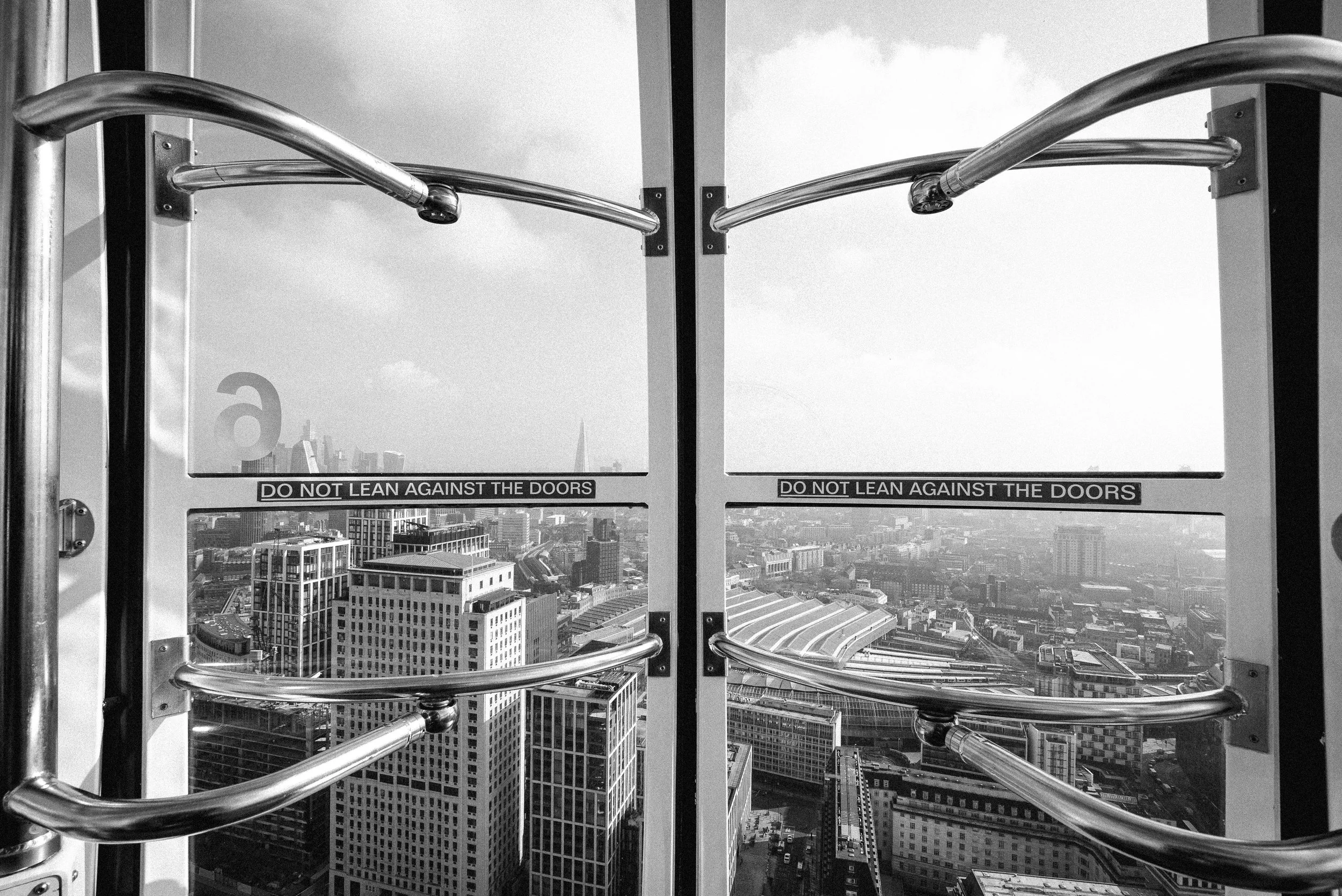 Black and white photo of a city skyline viewed through glass doors with metal railings and safety signs that say 'Do not lean against the doors'.