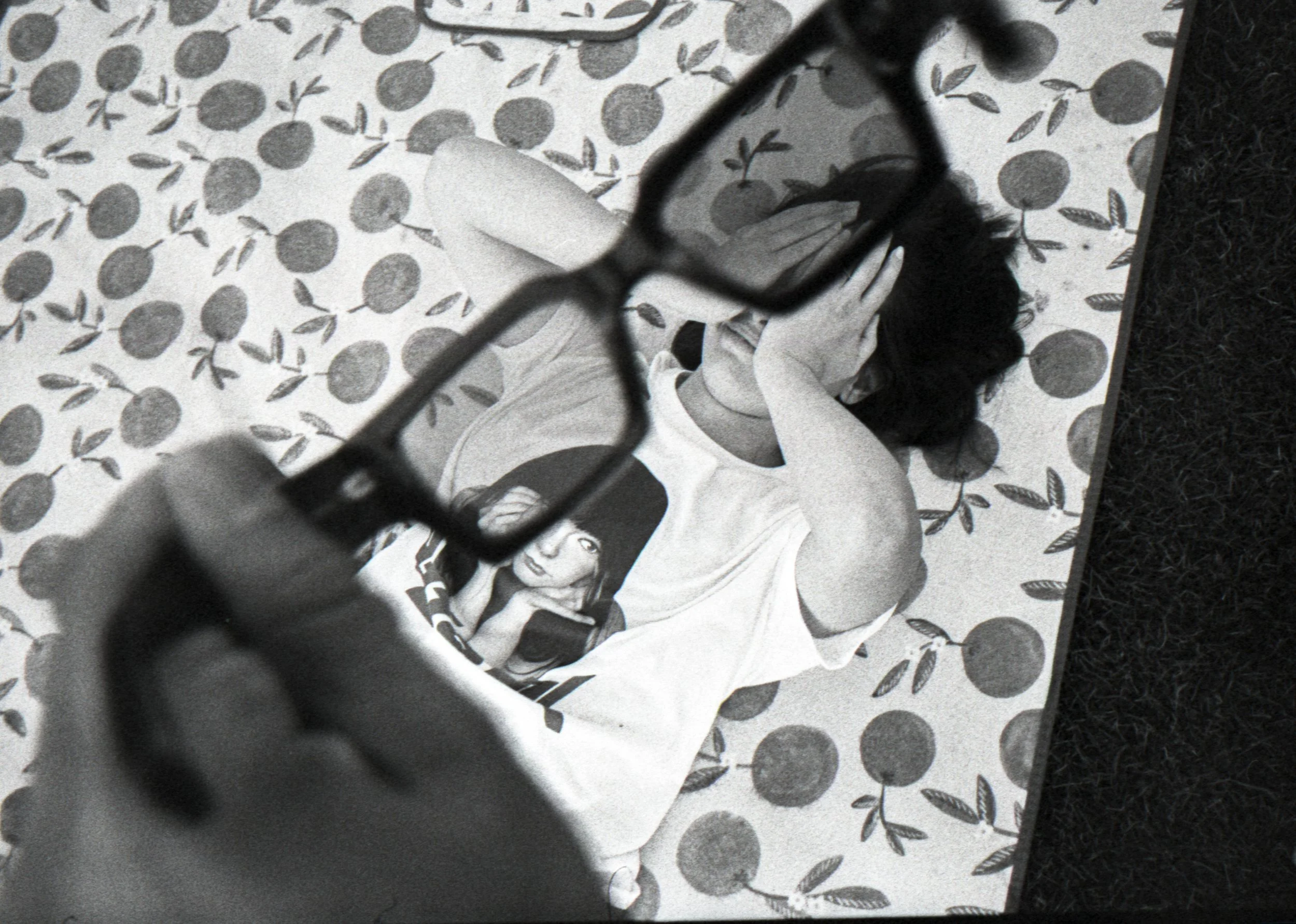 An overhead view of a person sitting on a patterned outdoor or patio rug with sunglasses and facing down next to a person lying on the rug, with a chain-link fence in the foreground.