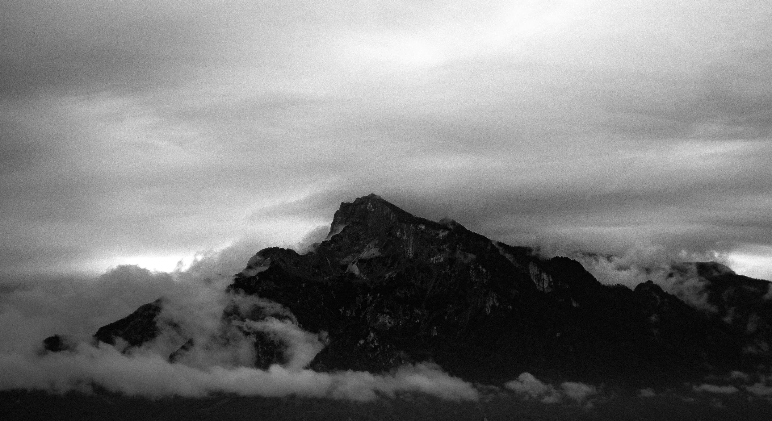 A black and white photograph of a mountain peak surrounded by mist and clouds with a dramatic, overcast sky.