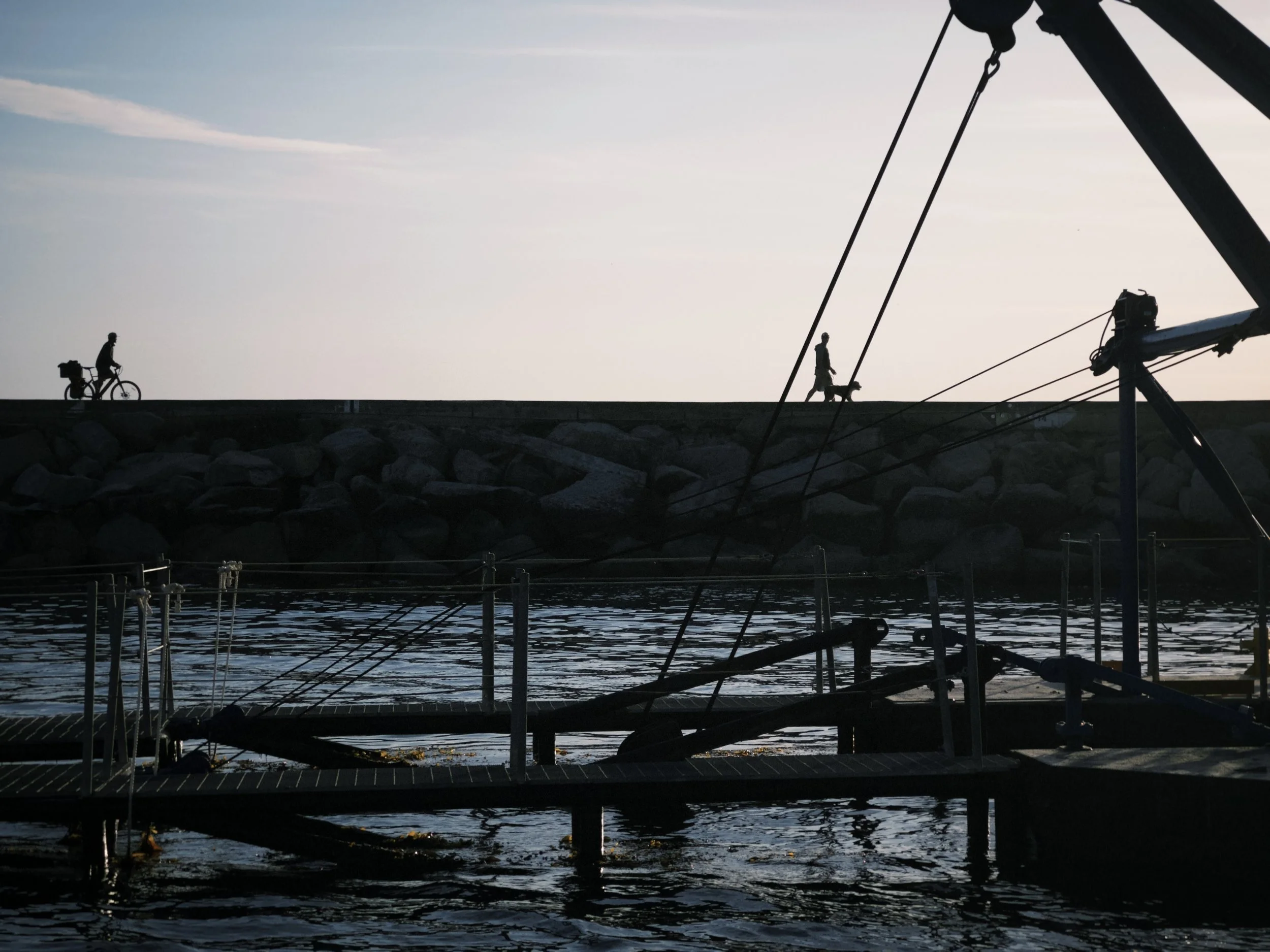 Silhouettes of two people walking and biking along a pier at sunset with boat rigging in the foreground.