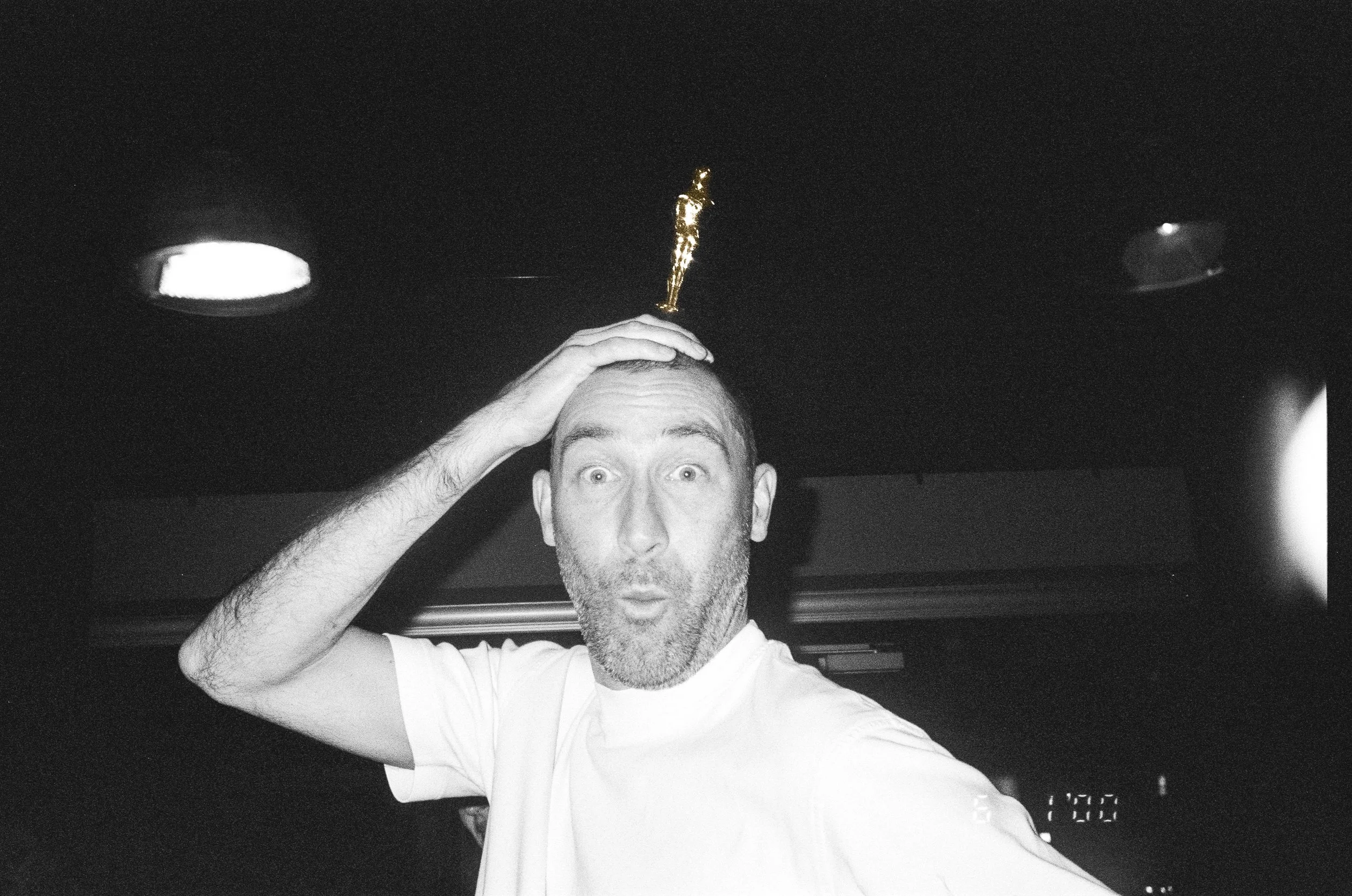 A man with short hair and a beard is holding his head with one hand, looking surprised or excited, with his mouth open. He is inside a vehicle, possibly a car, with a dark interior and ceiling lights.