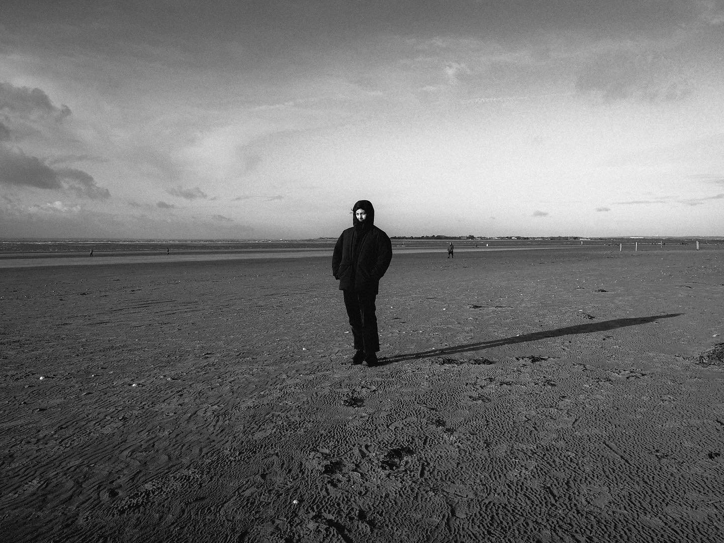 A person dressed in dark clothing standing on a sandy beach with a long shadow, with a few other people visible in the distance and an overcast sky.