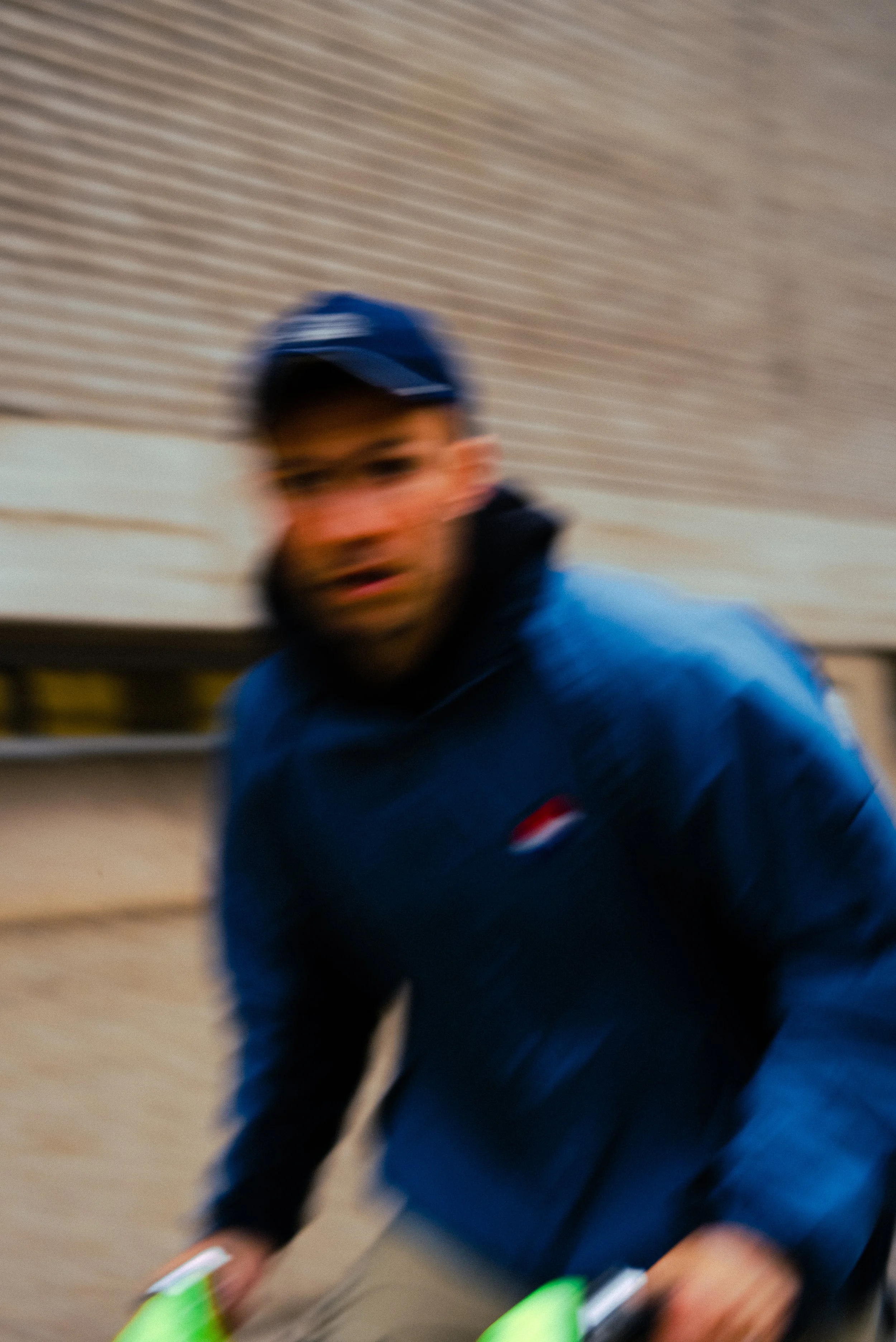A blurred photo of a man wearing a blue cap and jacket, in motion indoors, reaching towards the camera.