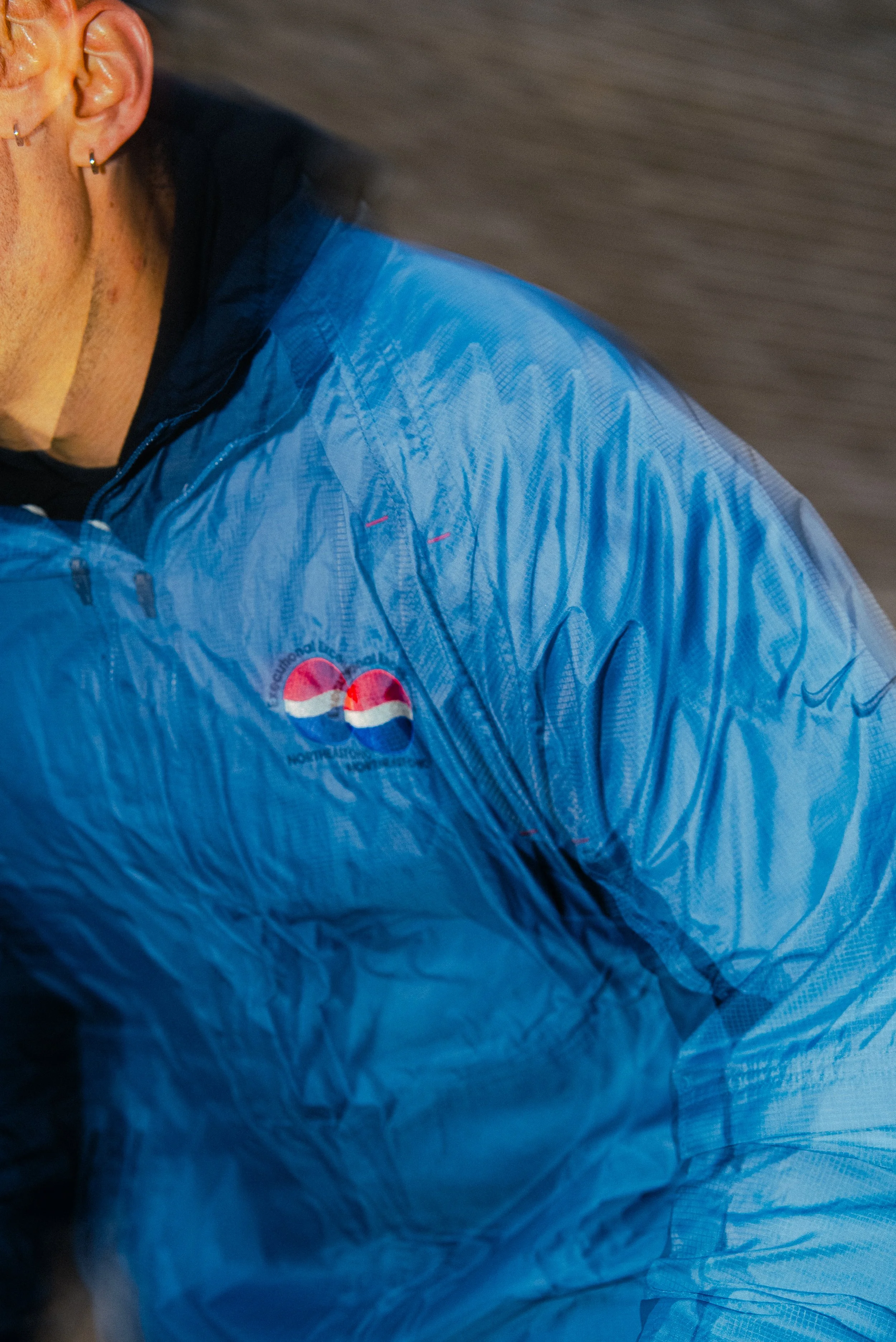 Close-up of a person wearing a blue jacket with a Pepsi logo, showing part of the face, ear with multiple earrings, and a portion of the background.