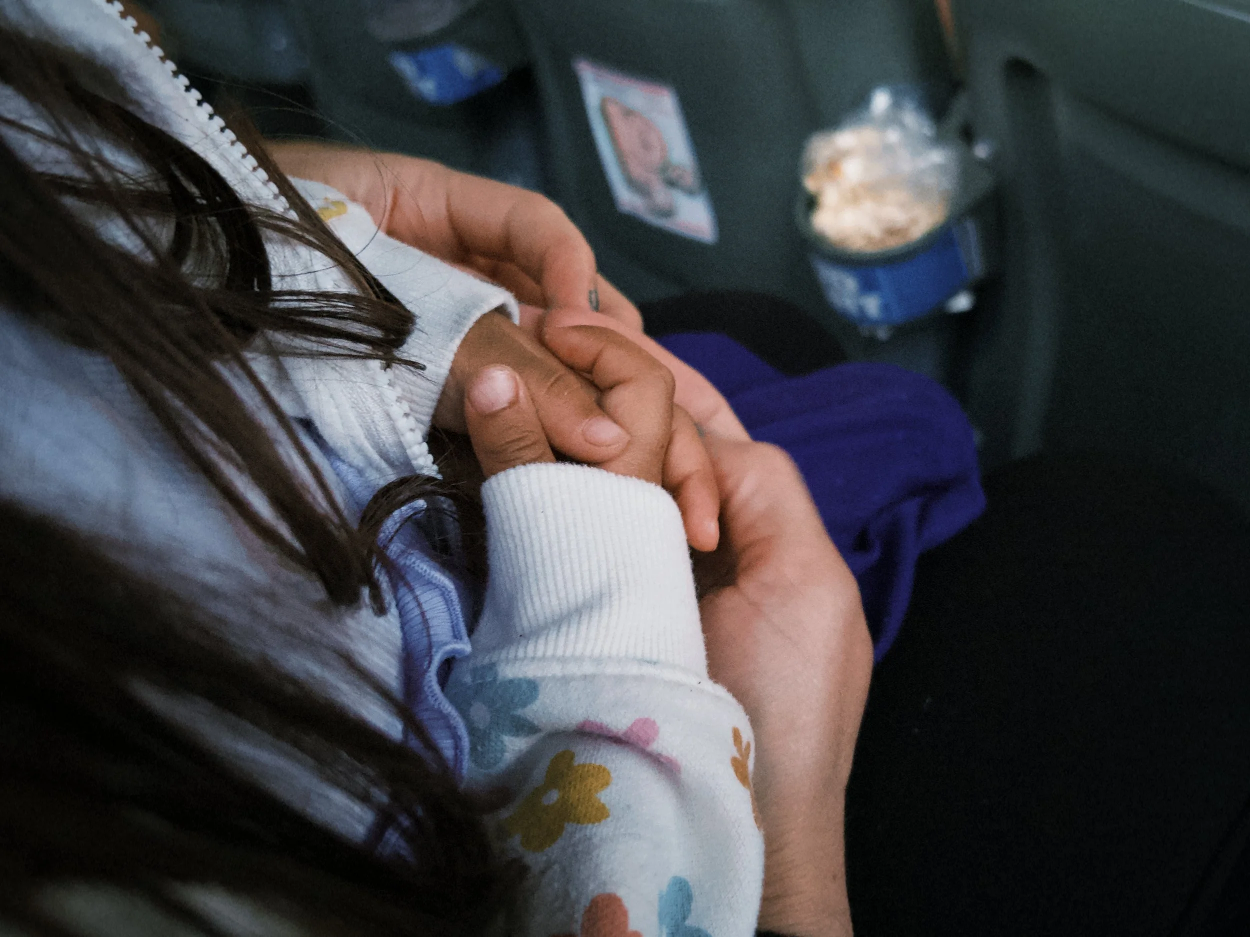 A person holding a child's hand in a vehicle, with the child's arm and part of their shirt visible. The person has long dark hair.