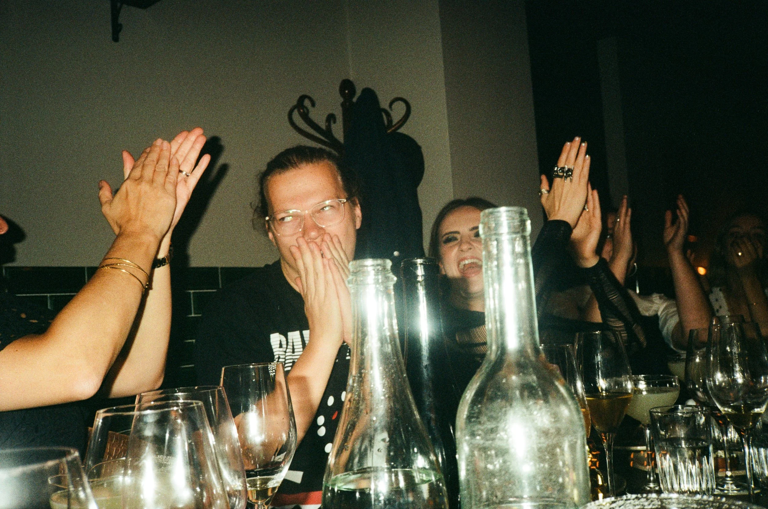 People at a social gathering or party raising hands in celebration, with drinks and glasses on the table, in a dimly lit setting.