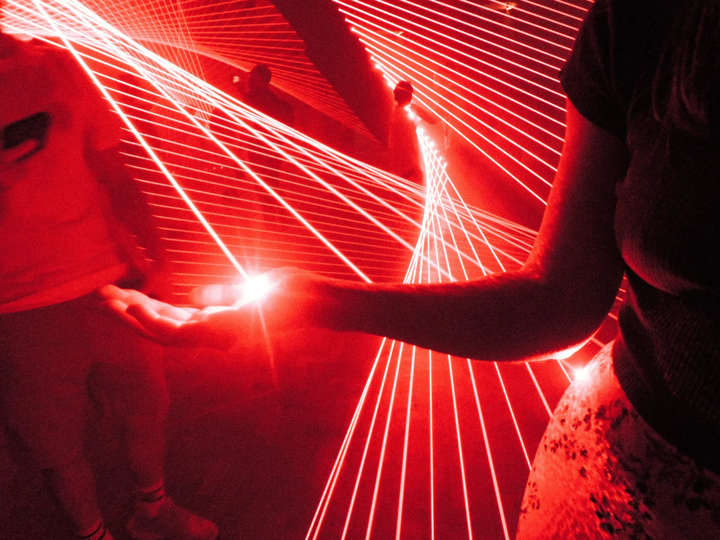 People standing and sitting around a red laser light installation creating geometric patterns on the floor and walls in a dark room.