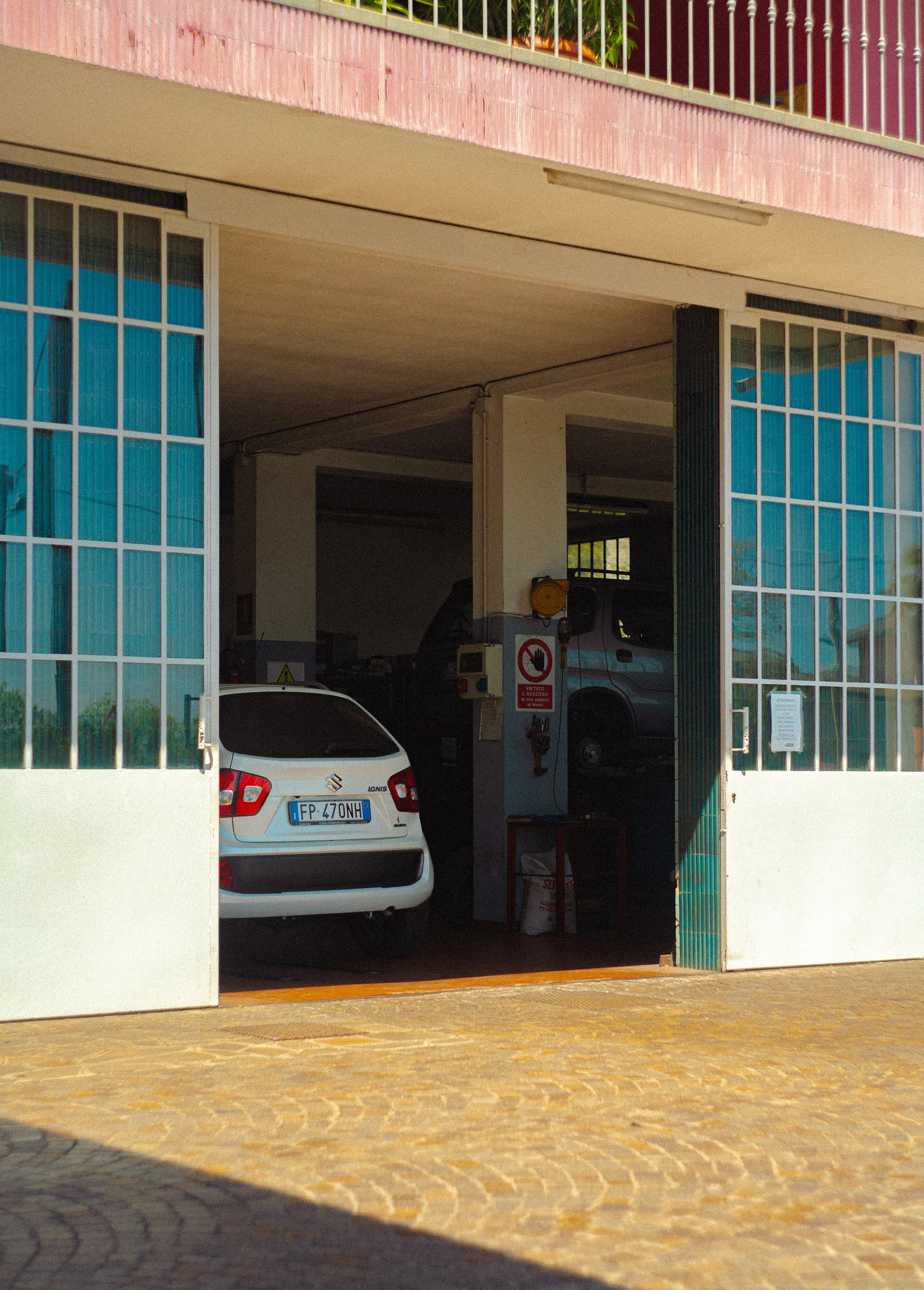 View of a garage with a white car parked inside and a van inside behind it. The garage has a large open door, with glass windows on either side. The exterior ground is paved with bricks, and a shadow covers part of it.