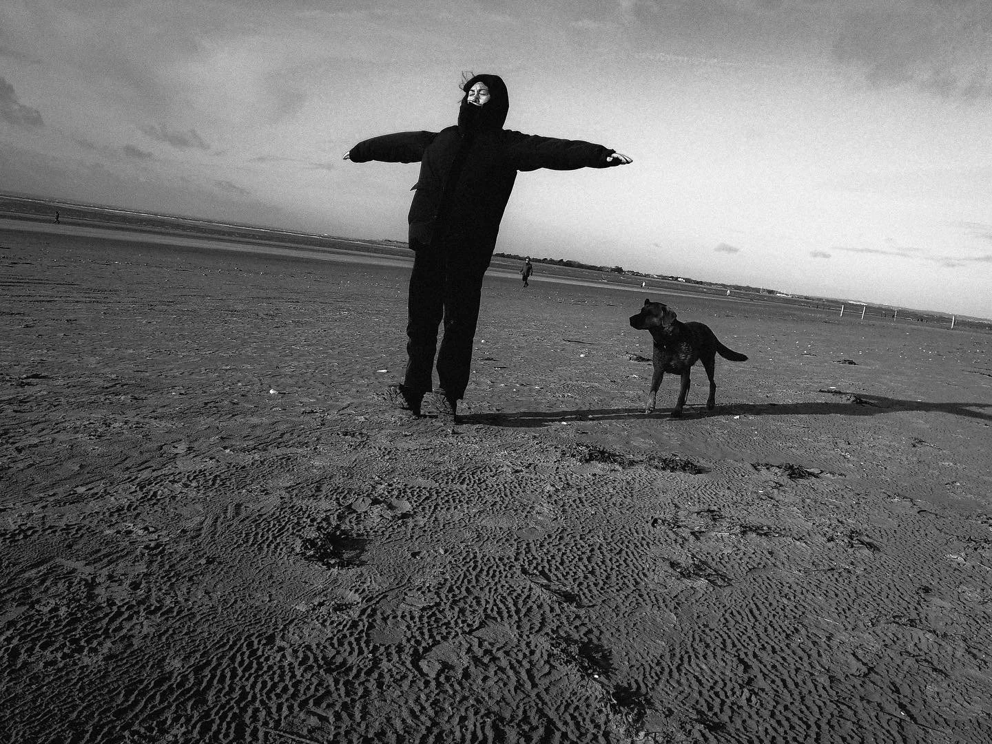 A person wearing a mask and dark clothing standing on a beach with arms outstretched next to a dog.