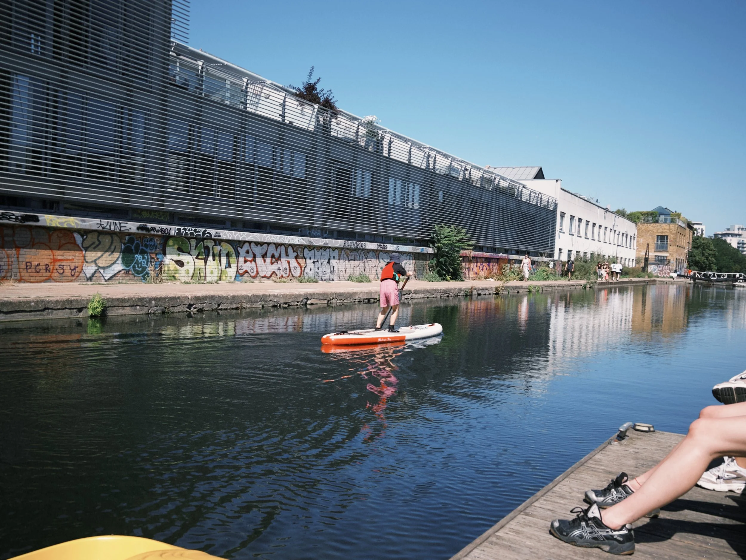 People sitting on a dock beside a canal, with one person paddleboarding on the water, in front of a modern building with graffiti on its lower wall, under a clear blue sky.