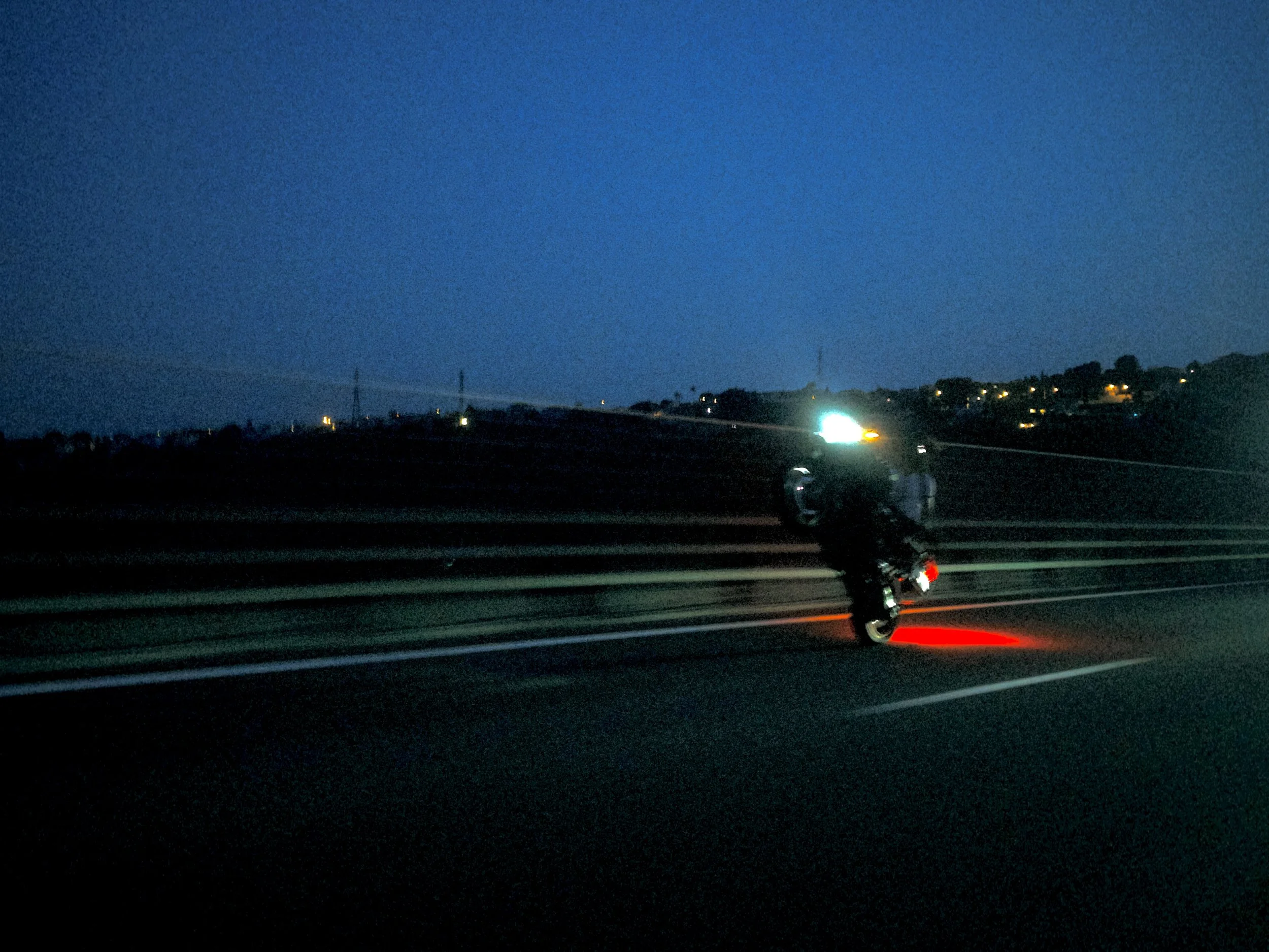 Nighttime view of a road with vehicles, city lights in the distance, and a clear dark blue sky.