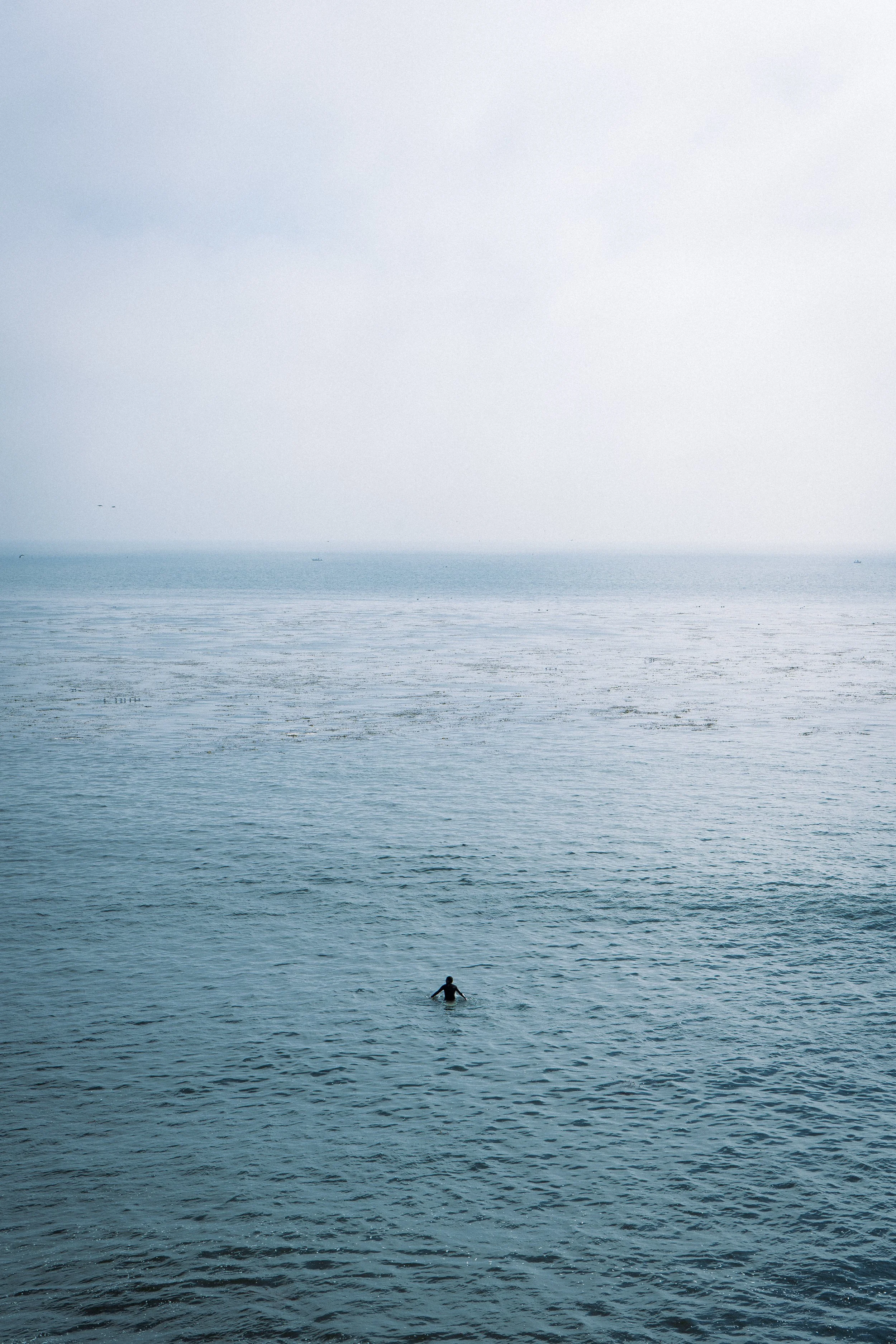 A person swimming alone in the ocean with a few boats in the distant horizon under a pale sky.