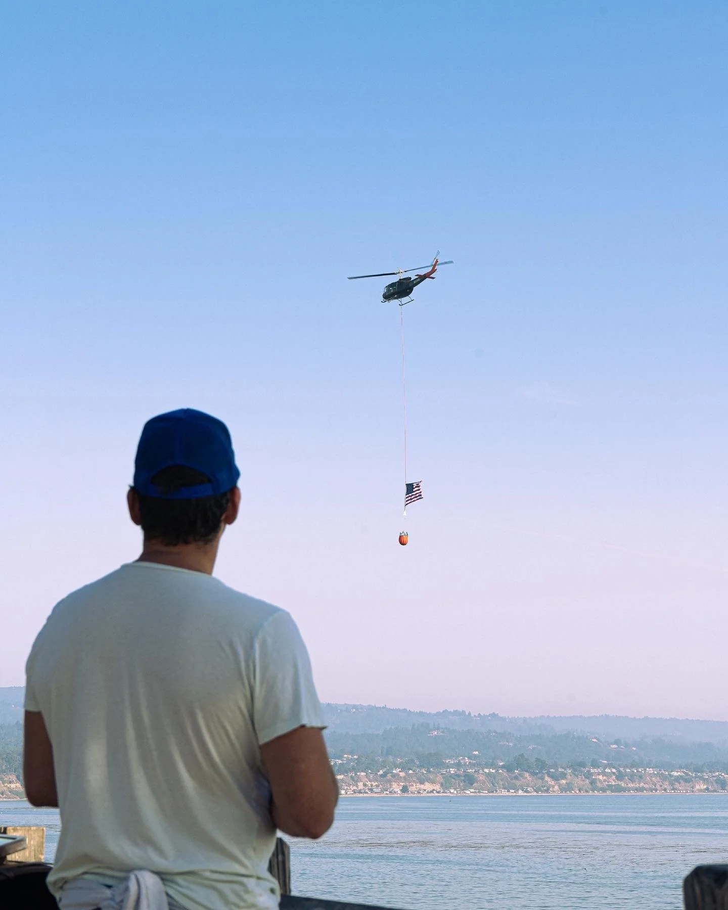 A man wearing a white t-shirt and a blue baseball cap is standing outdoors near a body of water, observing a helicopter in the sky. The helicopter is towing a line with an American flag and a bright orange float.