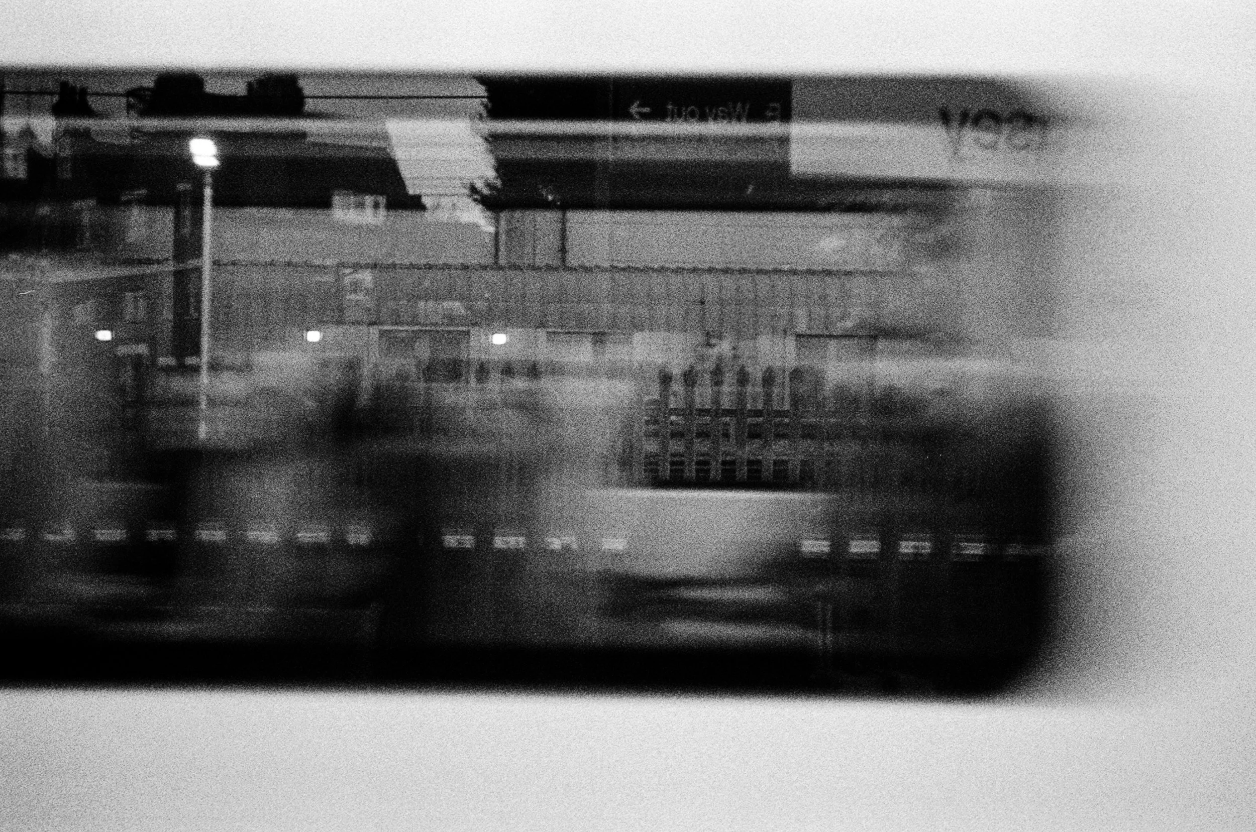 A black and white photo of a model train track with a train, with a sign in the background that reads 'E. WASH.' The train and tracks are blurry and the image has a grainy texture.