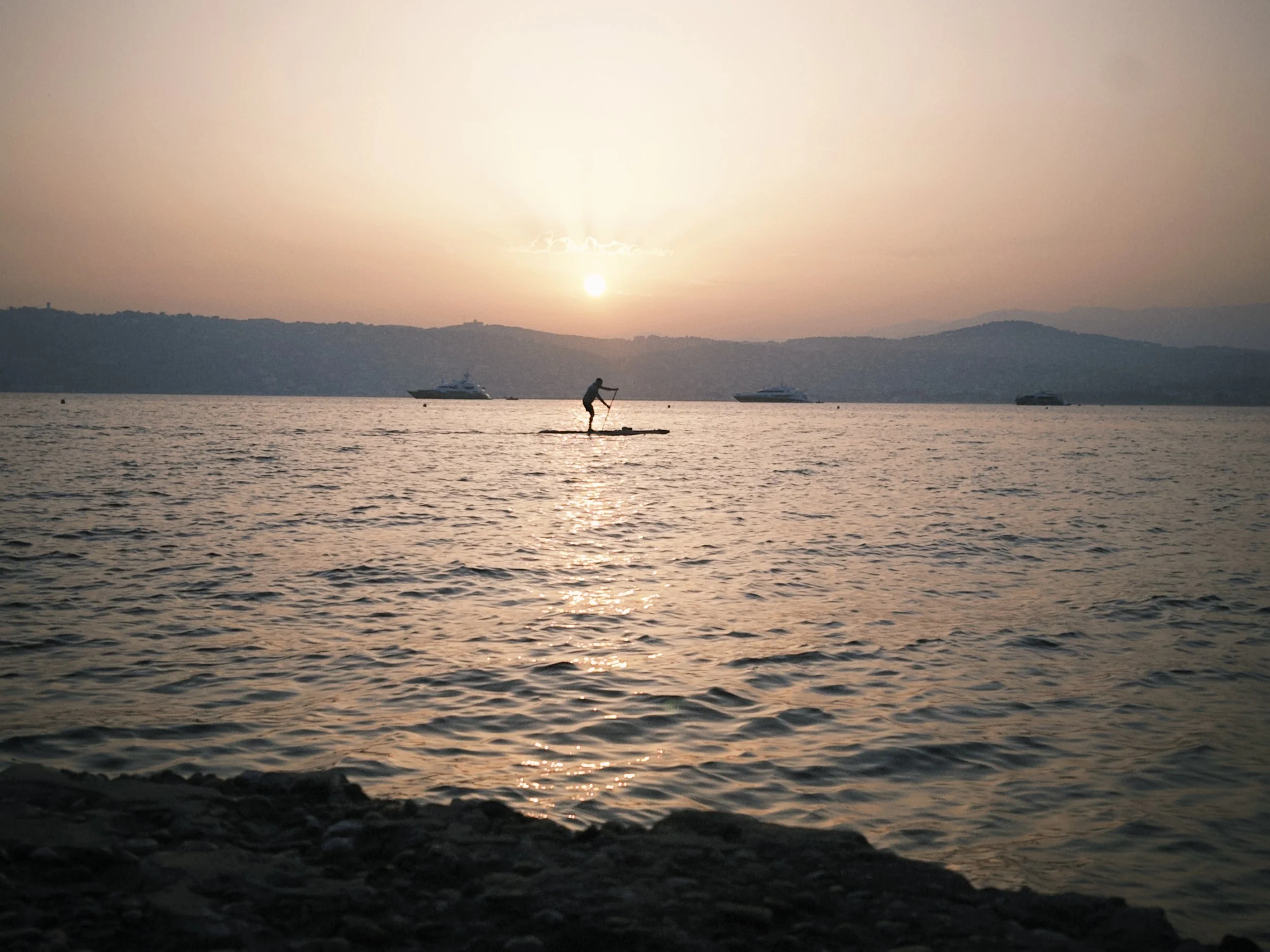 Person paddleboarding on a calm body of water during sunset, with boats anchored in the distance and hills on the horizon.