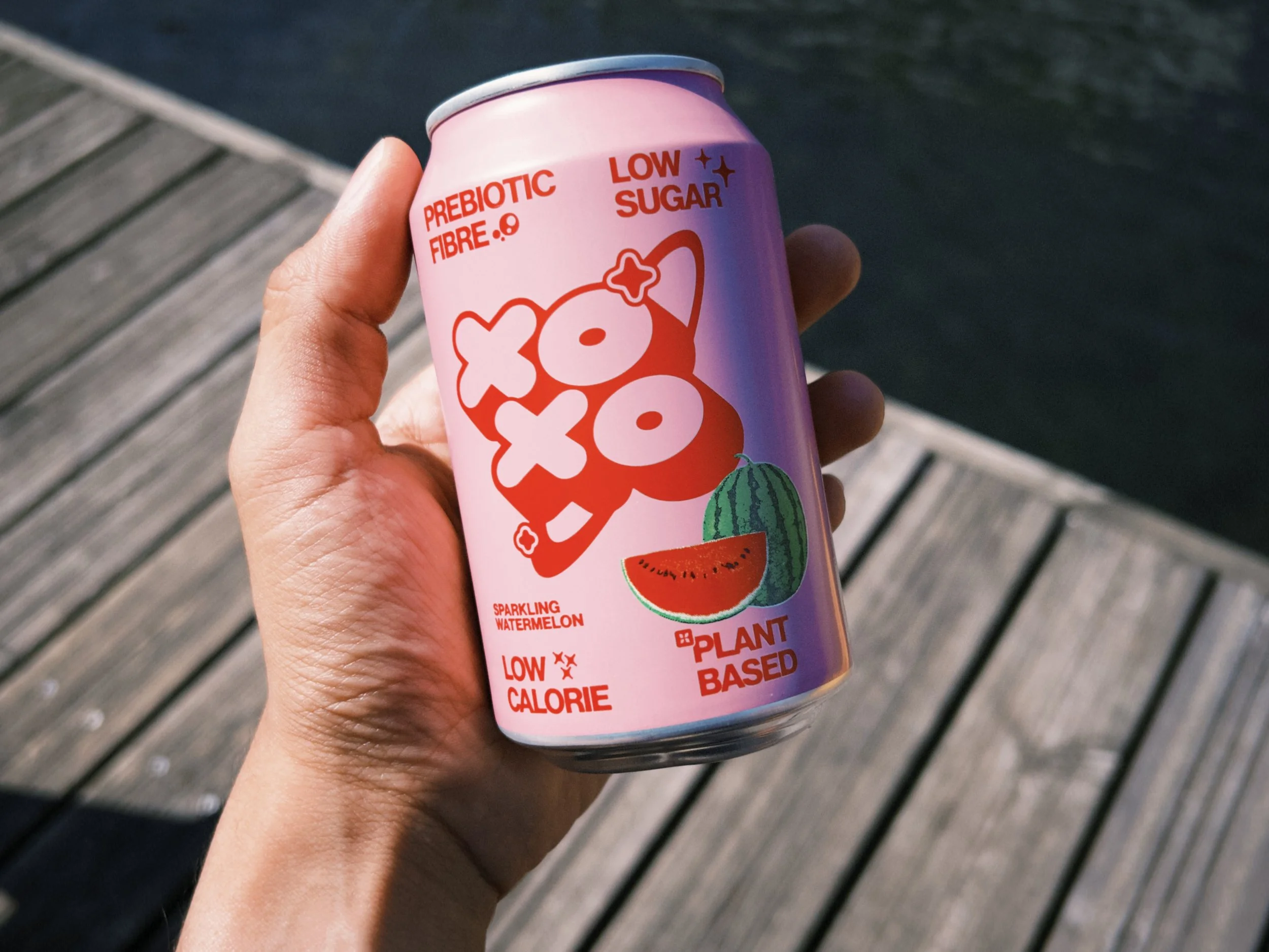 A hand holding a pink can of sparkling watermelon flavored beverage from Plant Based. The can features a watermelon illustration, the words "Prebiotic Fiber 0.8" and "Low Sugar" printed on it.
