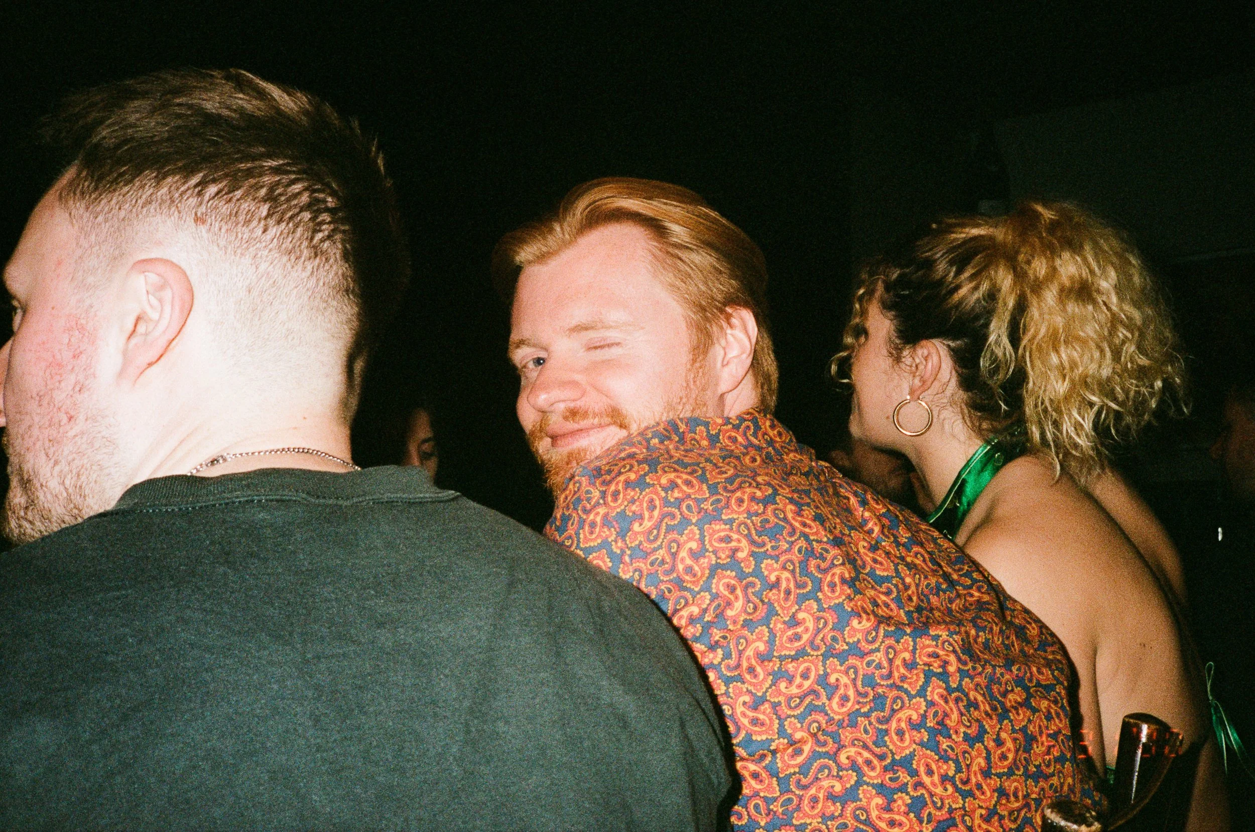 A group of people at a social gathering, with one man winking and smiling at the camera, wearing a patterned shirt, in a dimly lit setting.