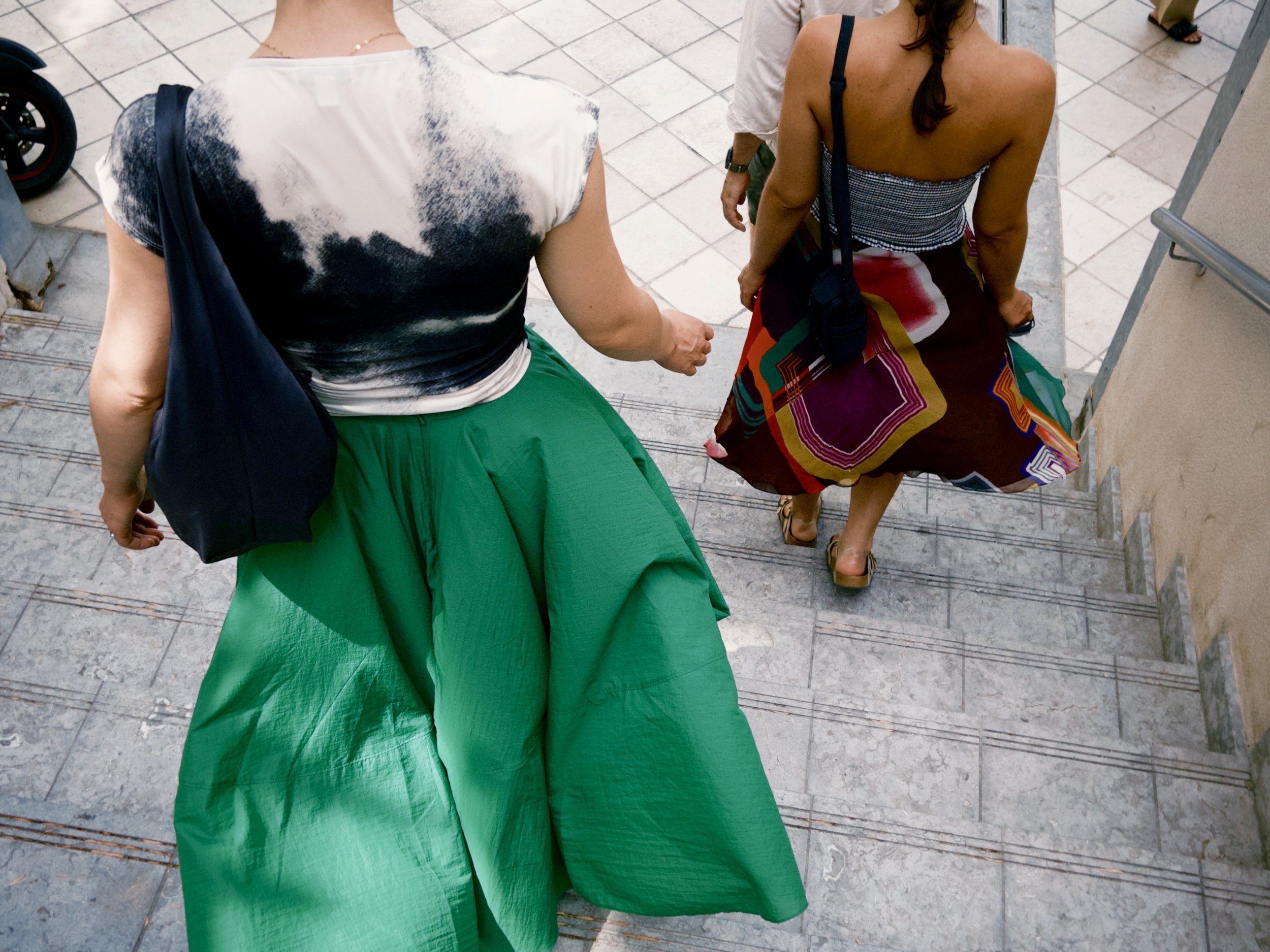 Top view of two women sitting on stairs, holding hands, with one wearing a black and white tie-dye shirt and a green skirt, and the other wearing a striped strapless top and colorful skirt.