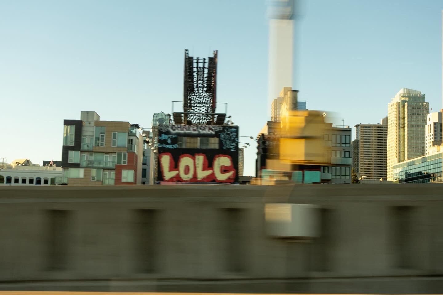 City skyline with tall buildings and a billboard that reads 'LOLC' in graffiti style, viewed from a moving vehicle.