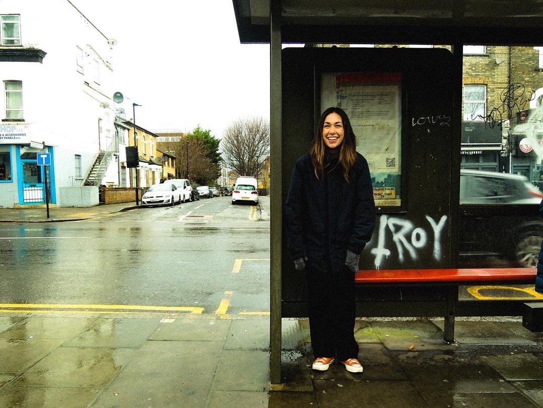 A woman with long brown hair, smiling, standing at a bus stop on a rainy day on an urban street with parked cars and buildings in the background.