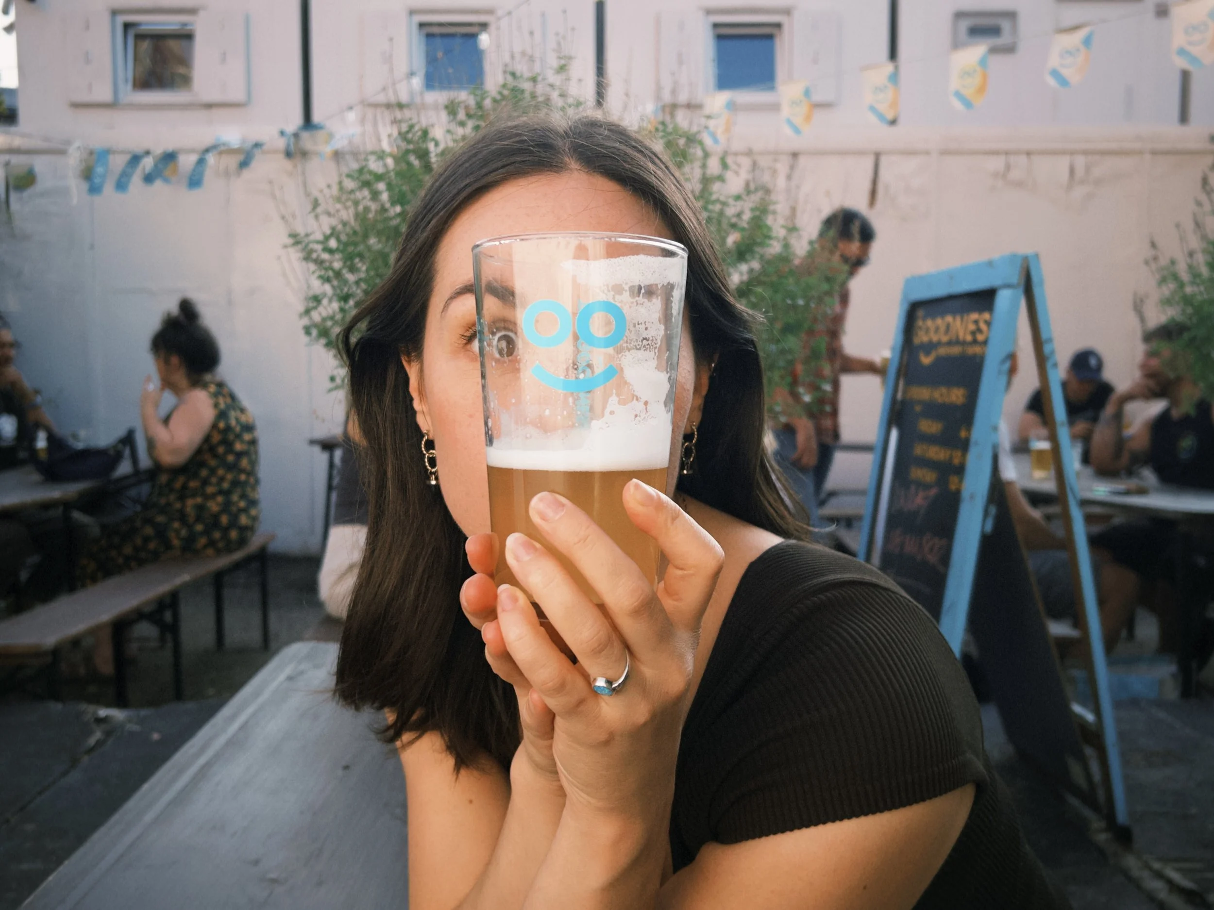 A woman holding a glass of beer with a smiley face logo in front of her face. In the background, there are people sitting at picnic tables and a man standing near a signboard, outdoors at a social gathering.