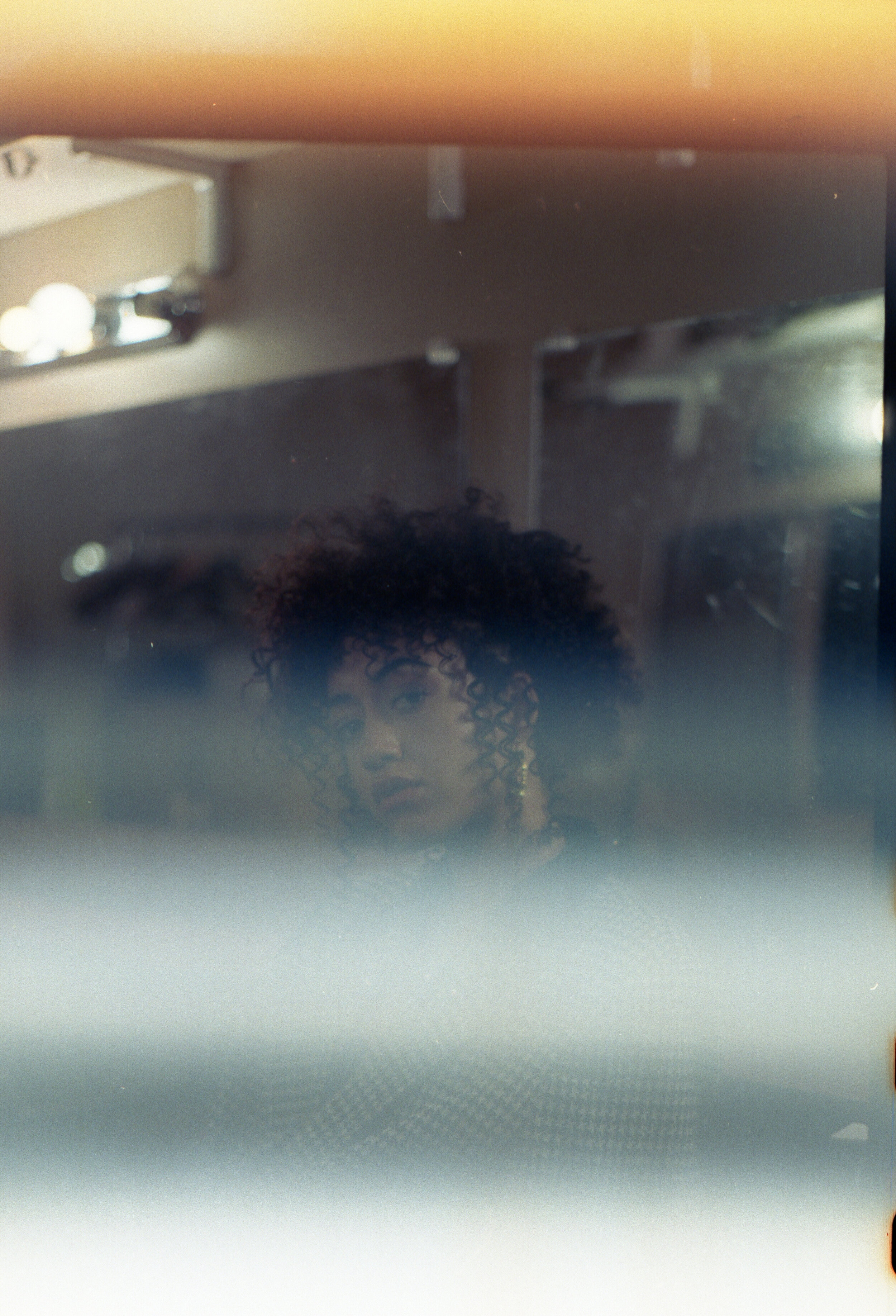 Reflection of a person with curly hair in a mirror or glass surface, with a warm light source above and a neutral background.