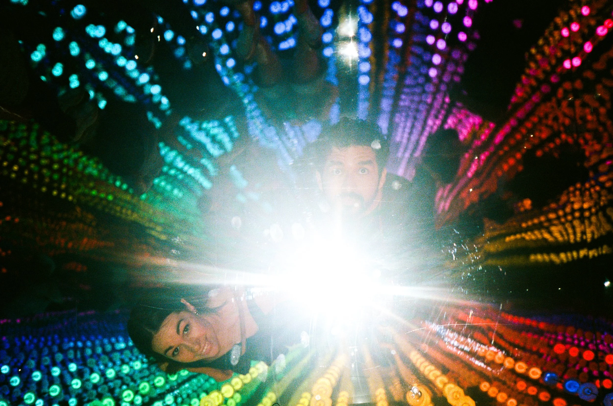 Two children smiling, surrounded by a vibrant rainbow-colored LED light installation in a dark space. One child is at the bottom left, and the other is in the center, looking upward. Bright light in the center casts a glow over the scene.