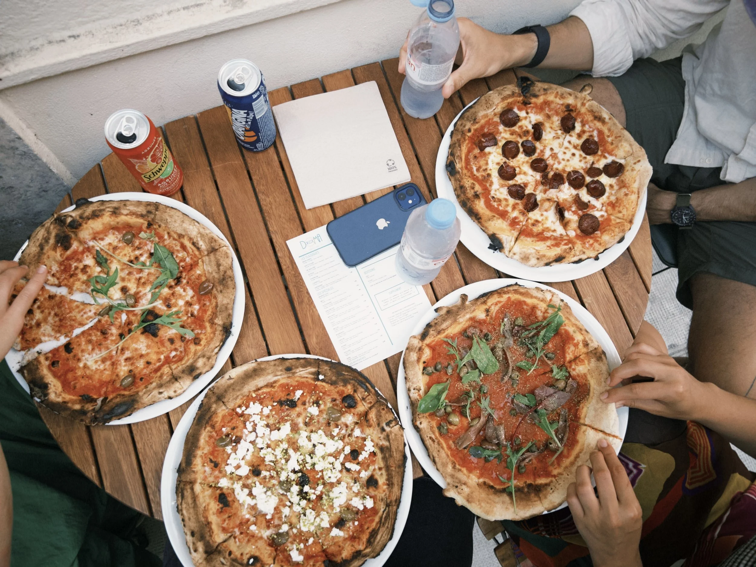 Multiple whole pizzas on plates with various toppings including pepperoni, herbs, and cheese, on a wooden table. People are reaching for slices, and there are drinks and a phone on the table.