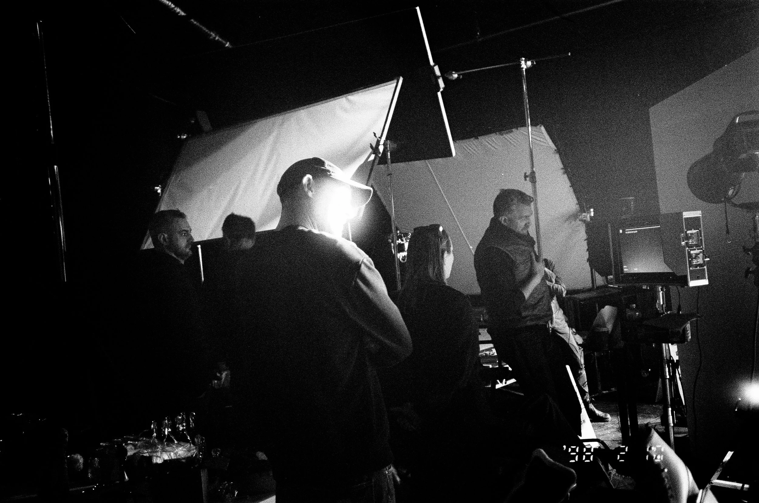 Black and white photo of a film or photography studio with multiple people working or preparing equipment, including large umbrellas and lighting fixtures.