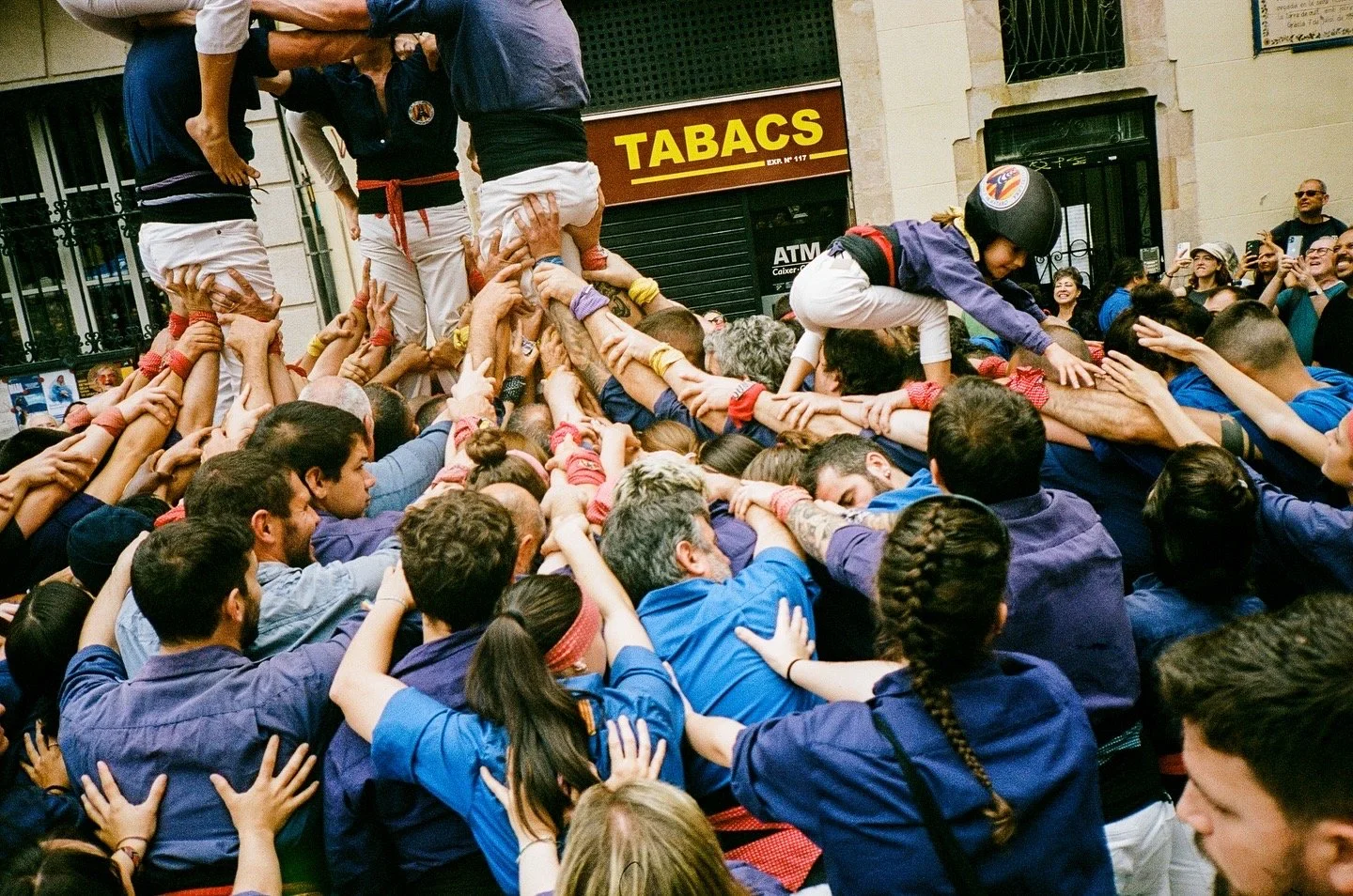 People participating in a human tower or 'castell', wearing traditional shirts and headbands, with onlookers taking photos in the background.