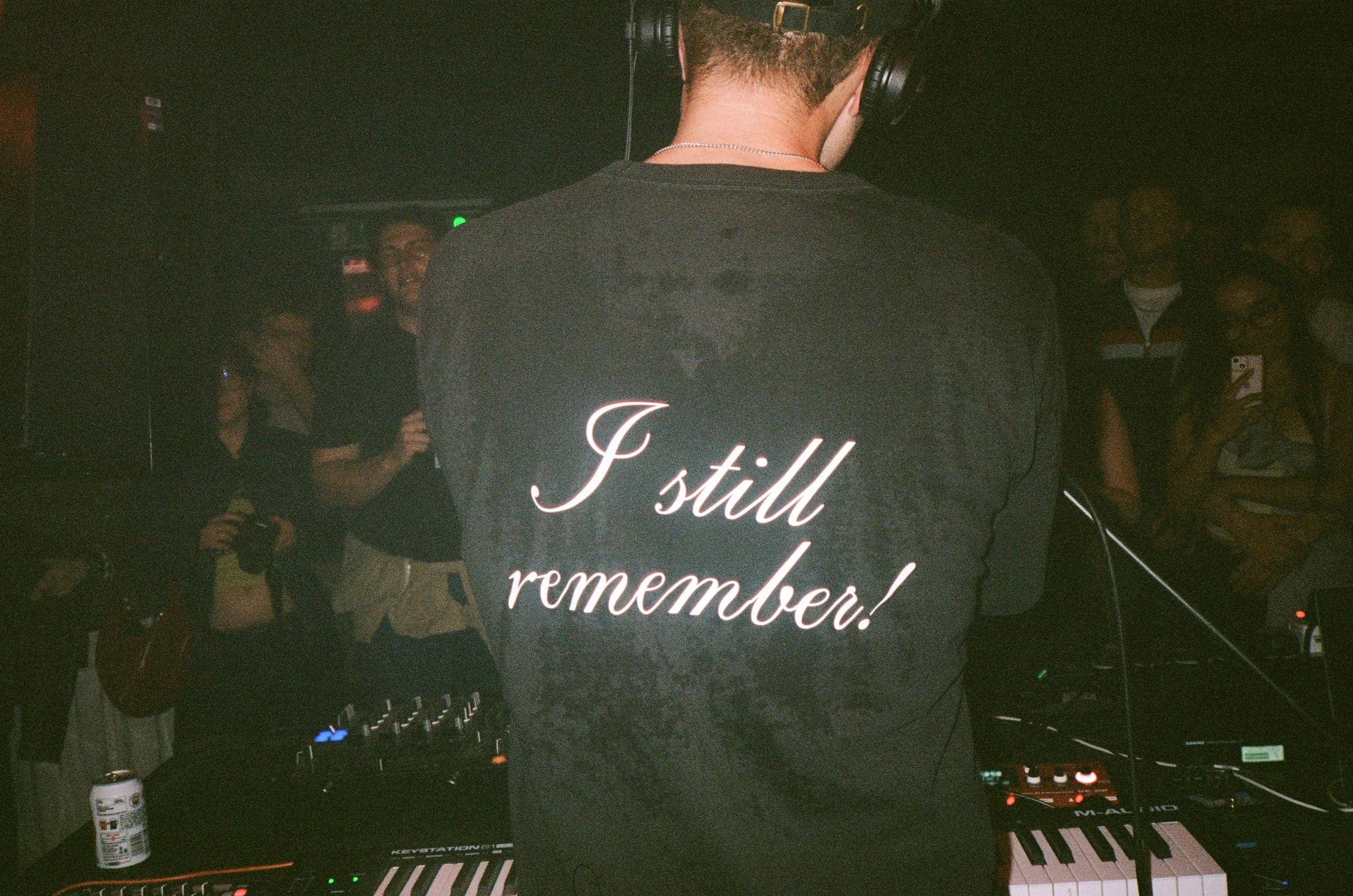 A person with short hair wearing headphones and a black T-shirt with white and pink lettering that says 'I still remember' is standing in front of DJ equipment. The image appears to be taken at a party or event with people in the background, some hol